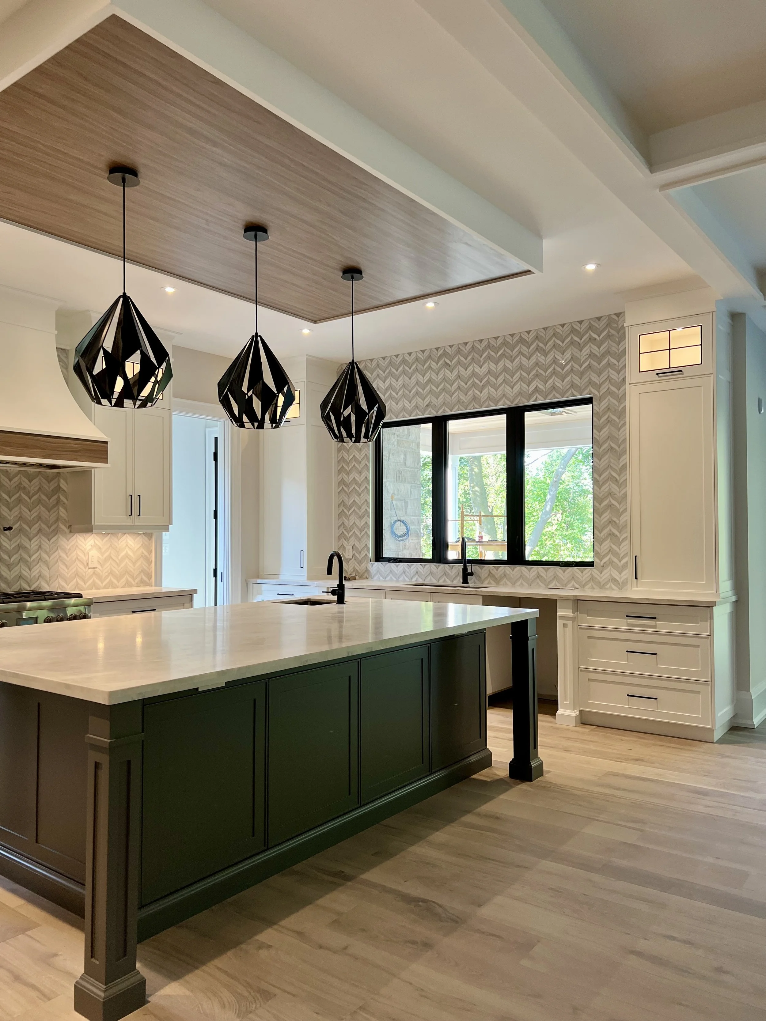 Modern kitchen with a large island with dark green cabinetry, three black pendant lights, white countertops, and a window overlooking greenery outside.