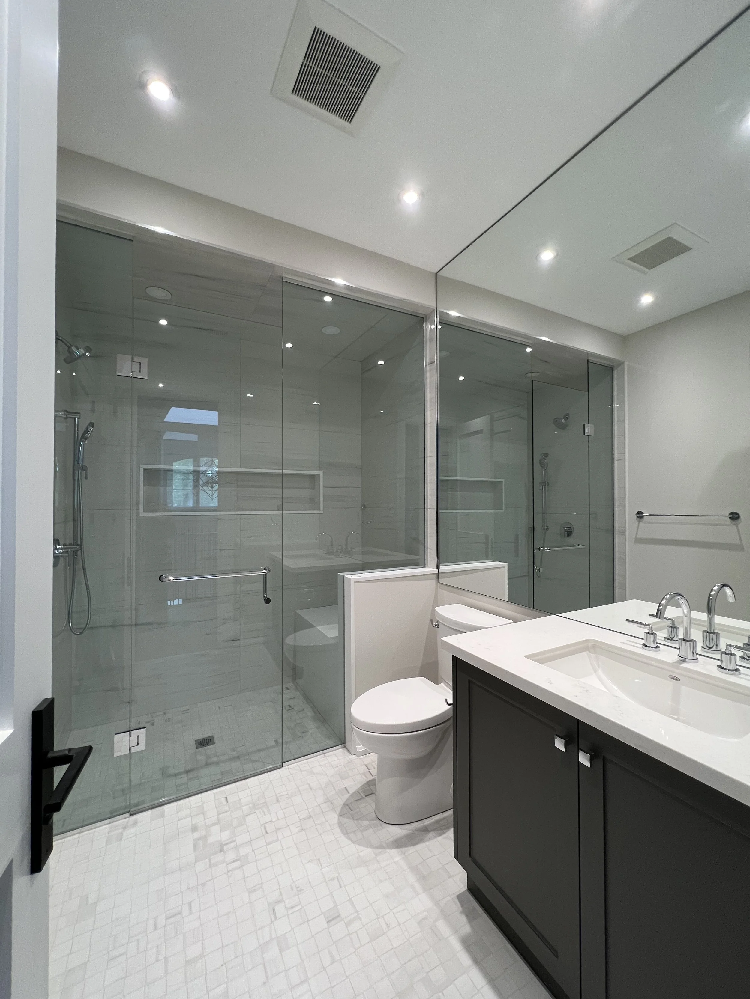 Modern bathroom with glass shower enclosure, white tiled floor, toilet, and gray vanity with white countertop and sinks.