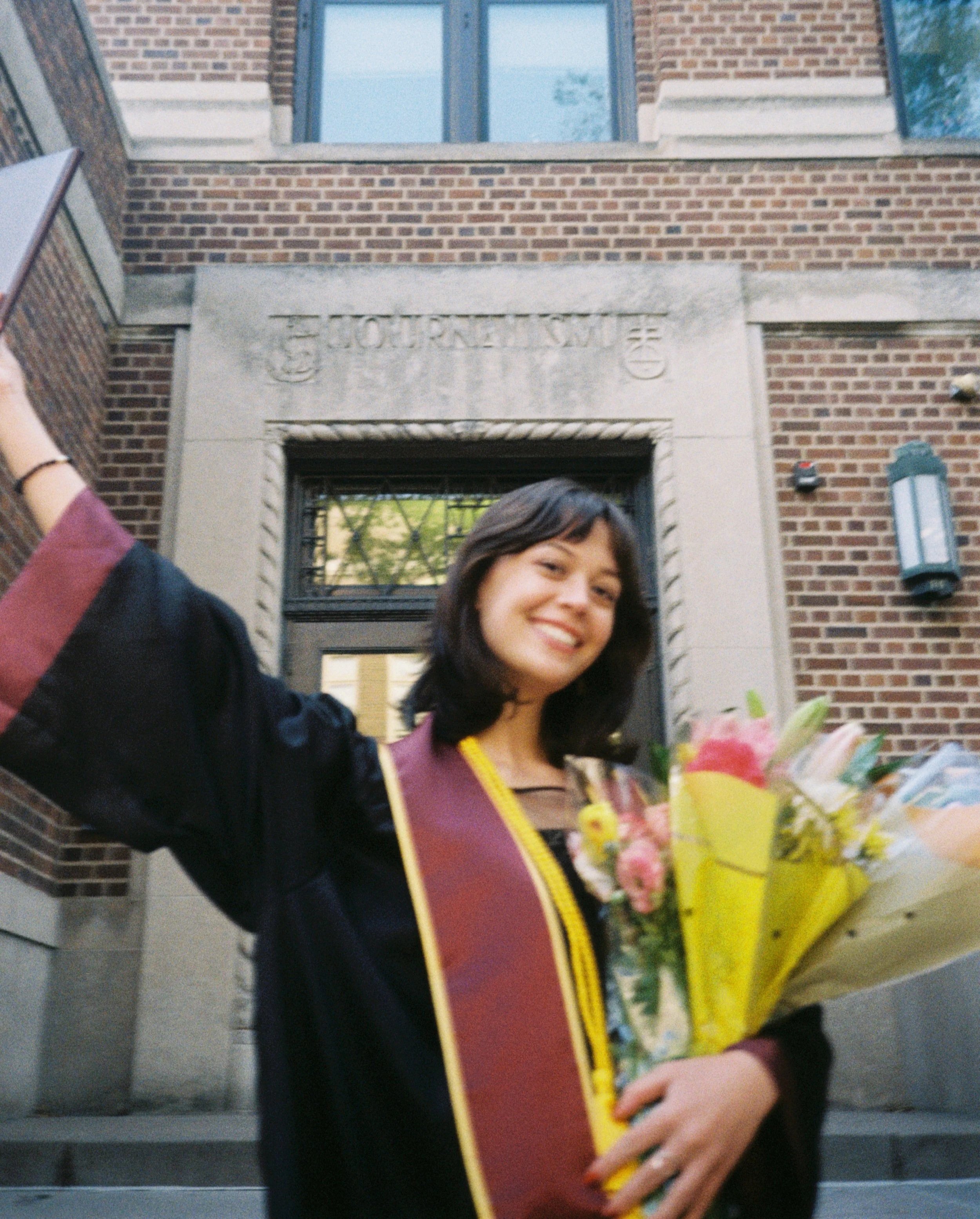 Sophia Marschall stands in a graduation gown holding flowers in front of a University of Minnesota building