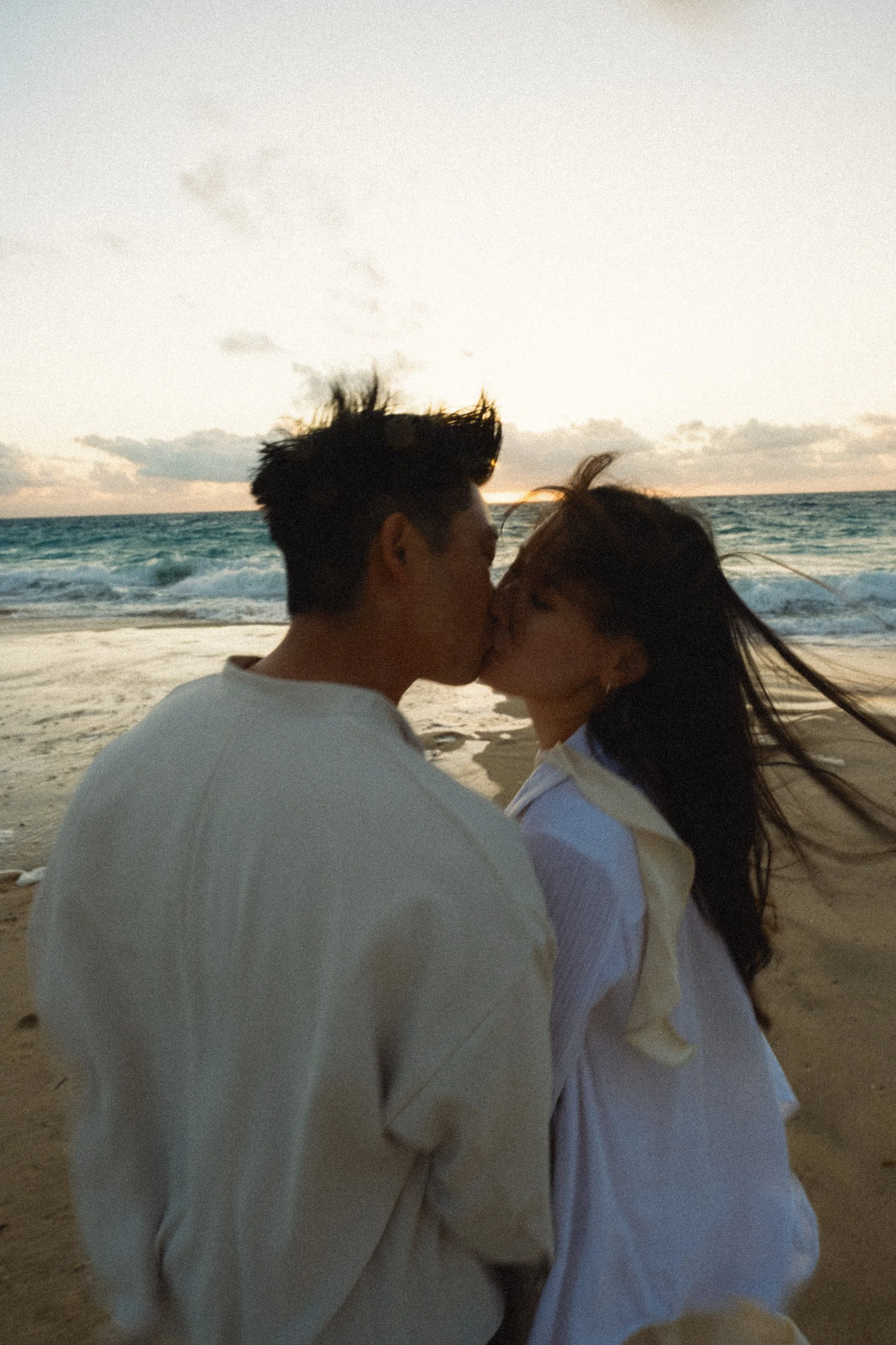 A couple kissing on the beach during sunset, with the ocean and sky in the background.