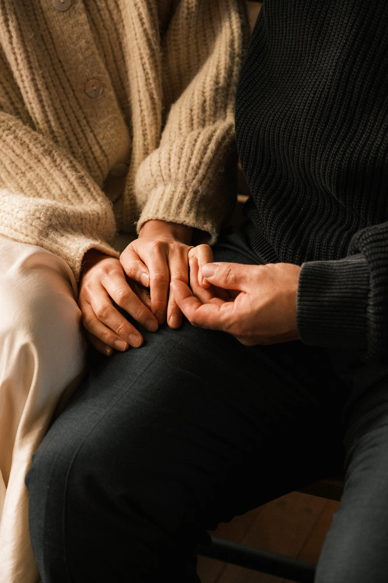 Two people holding hands, one resting their hand on the other's lap, both wearing cozy sweaters.