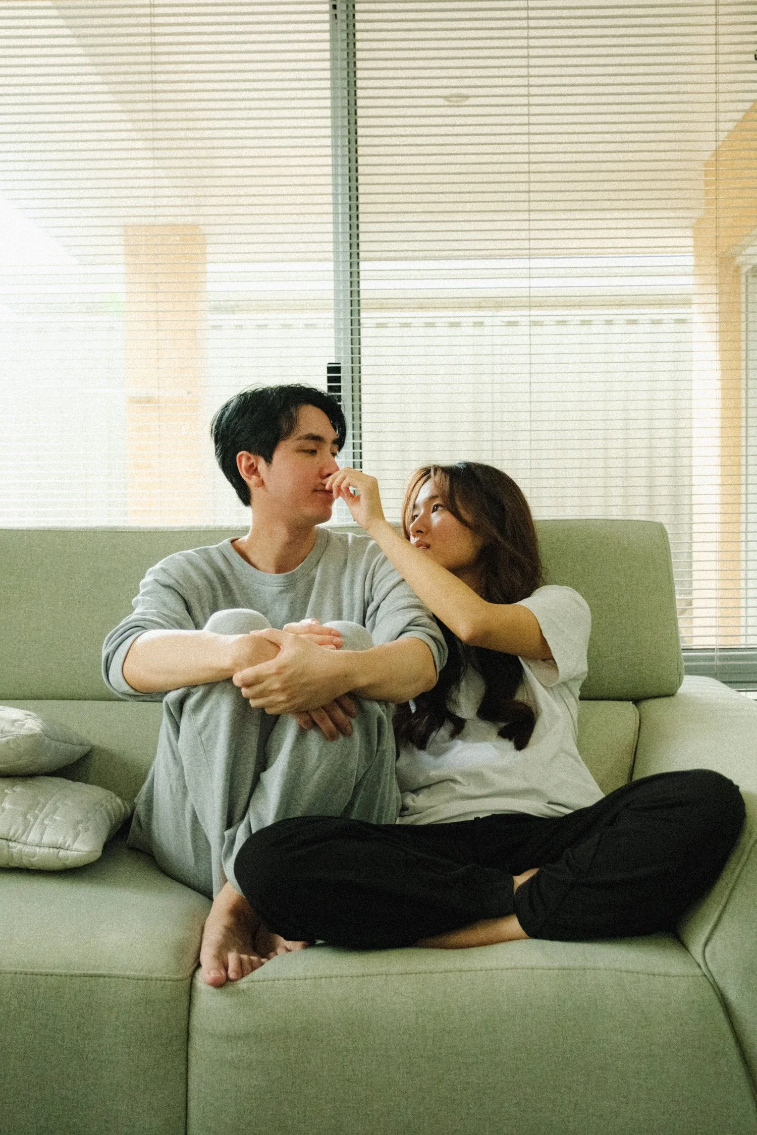 A young man and woman sit on a green sofa in front of large window blinds. The woman playfully touches the man's face as they look at each other affectionately. The man is dressed casually in a gray shirt and pants, and the woman is dressed in a white top and black pants. The room is softly lit with natural light coming through the window.