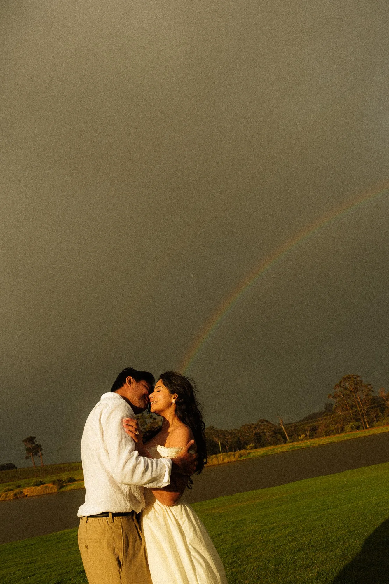 A couple embraces outdoors during sunset, with a rainbow visible in the sky and trees and a lake in the background.