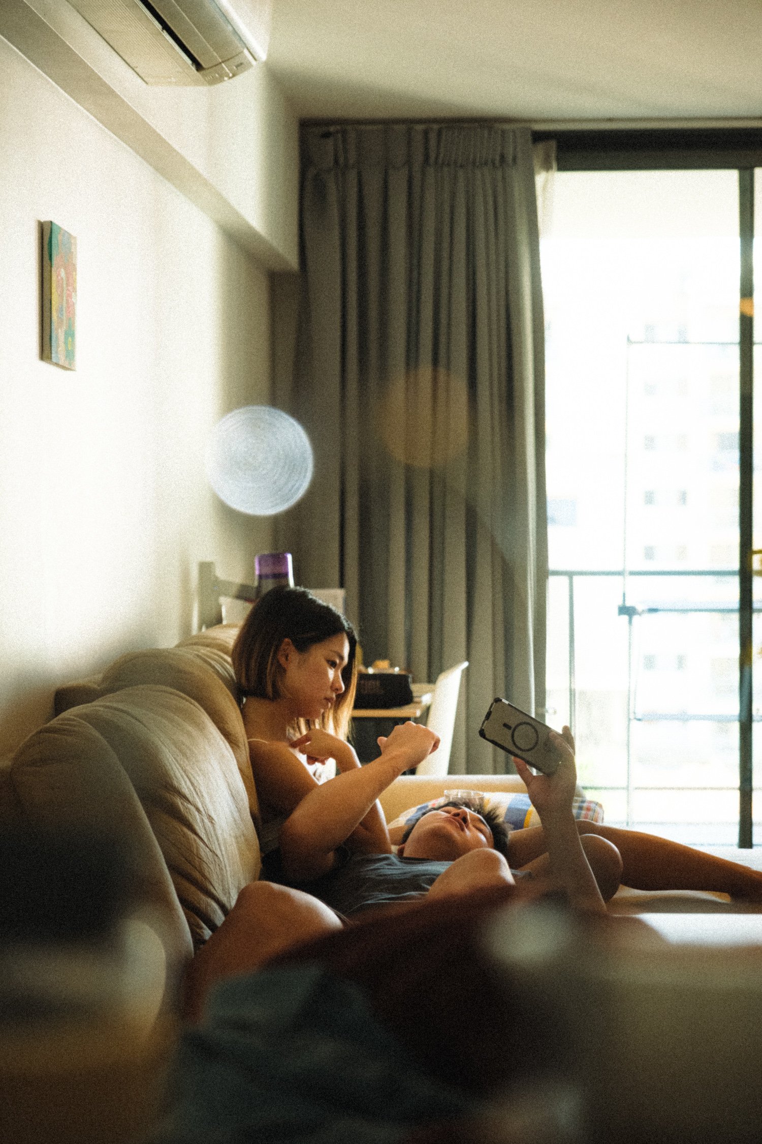 A woman and a man relaxing on a beige sofa inside a room with a balcony door, with the woman appearing to look at the man and the man lying down holding a phone.