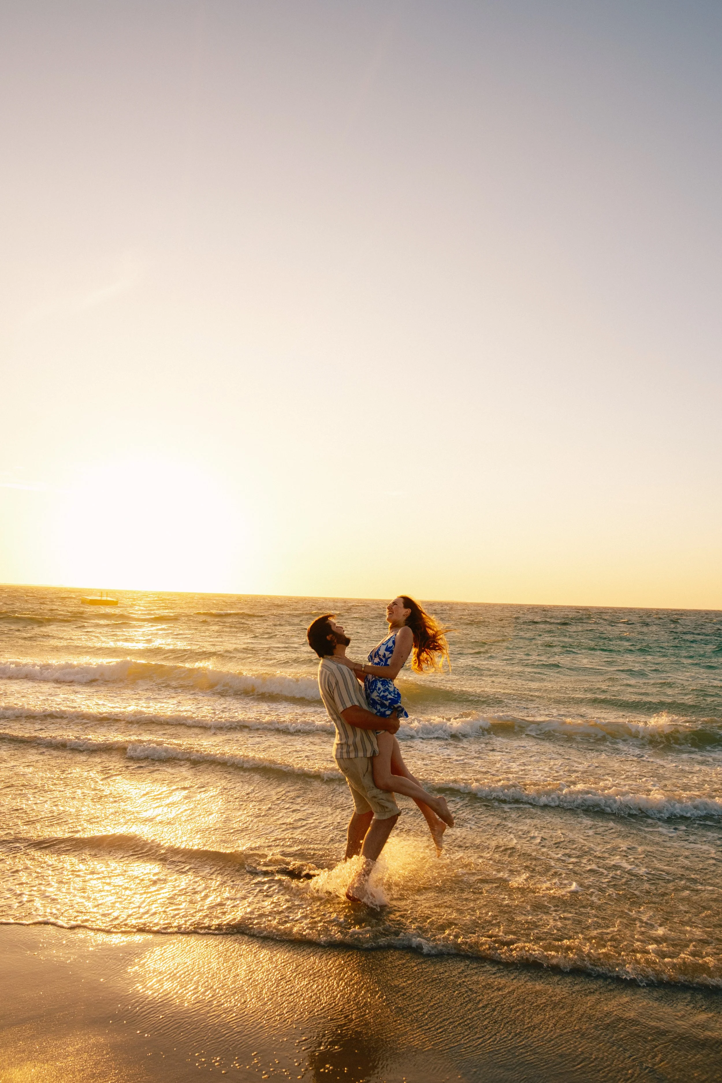 A couple at the beach during sunset. The man is holding the woman in his arms, and both are smiling and enjoying the moment. The sea and the setting sun are visible in the background.