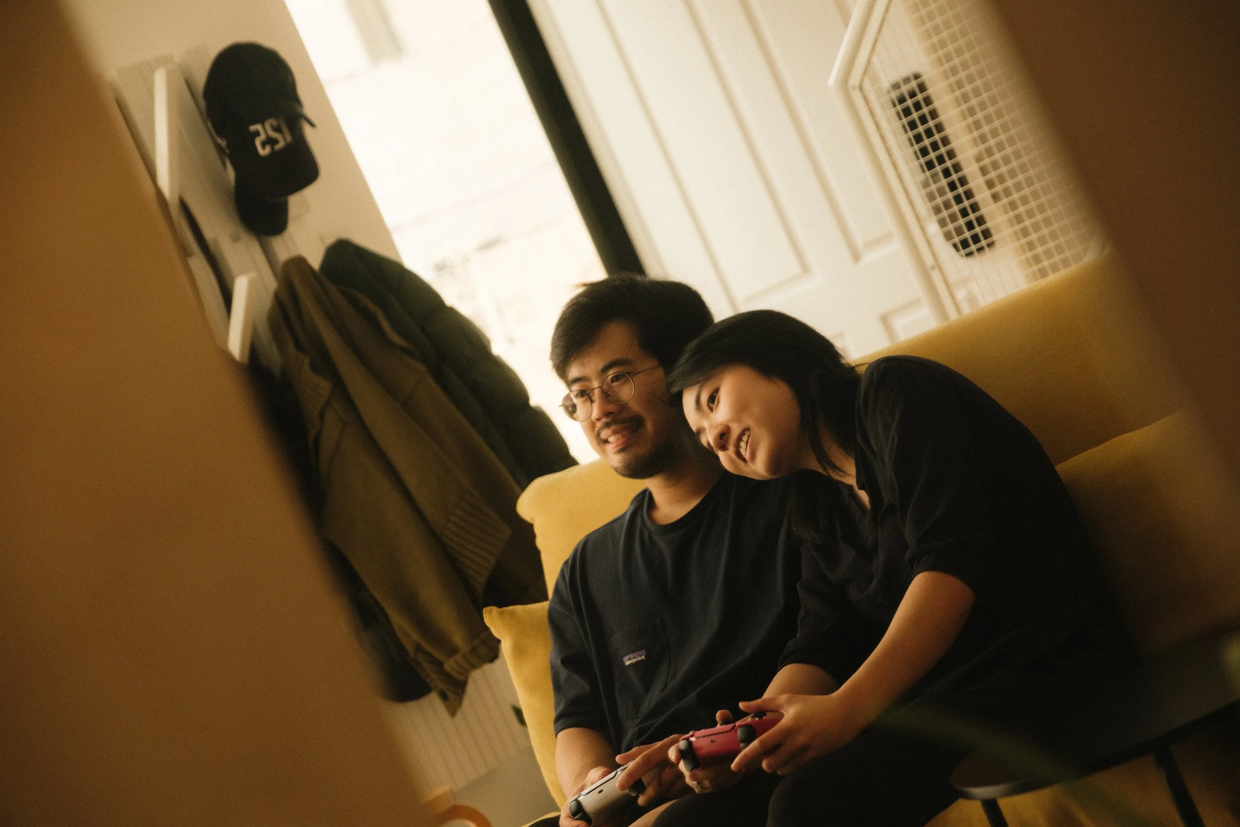 Two young people sitting on a yellow couch, one of them holding a Nintendo Switch gaming console, in a brightly lit room.