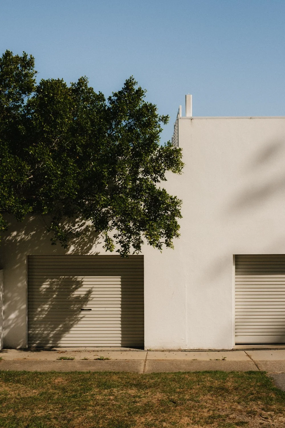 A white building with two garage doors, a tree casting shadows on the wall, and a clear blue sky.