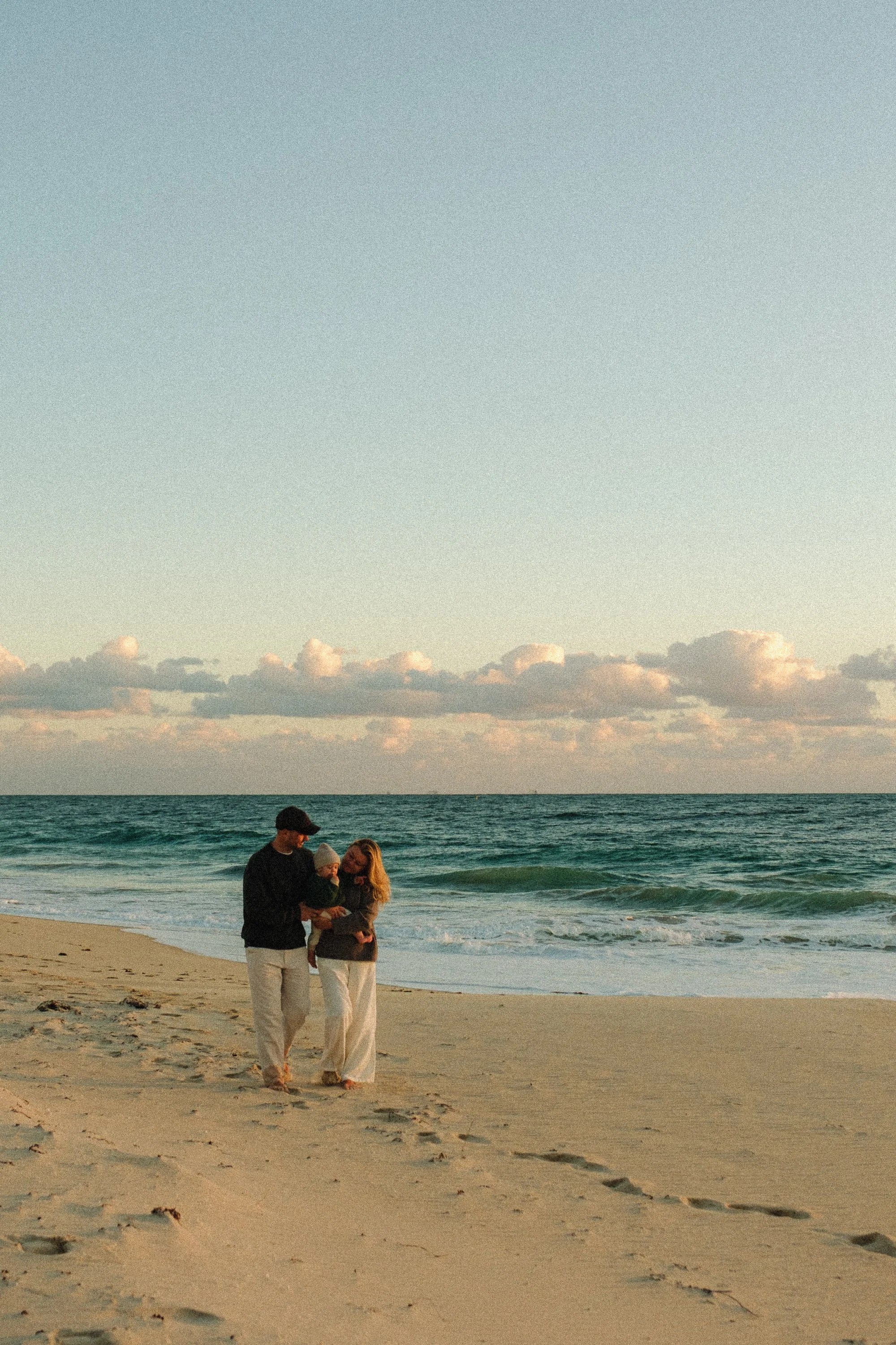 A family of three walking along the beach at sunset, with the ocean and clouds in the background.