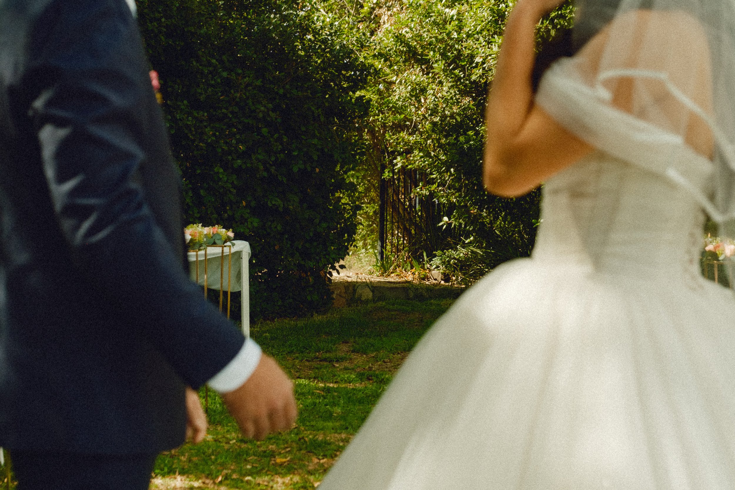 People holding hands at an outdoor wedding ceremony, with greenery and floral decorations in the background.