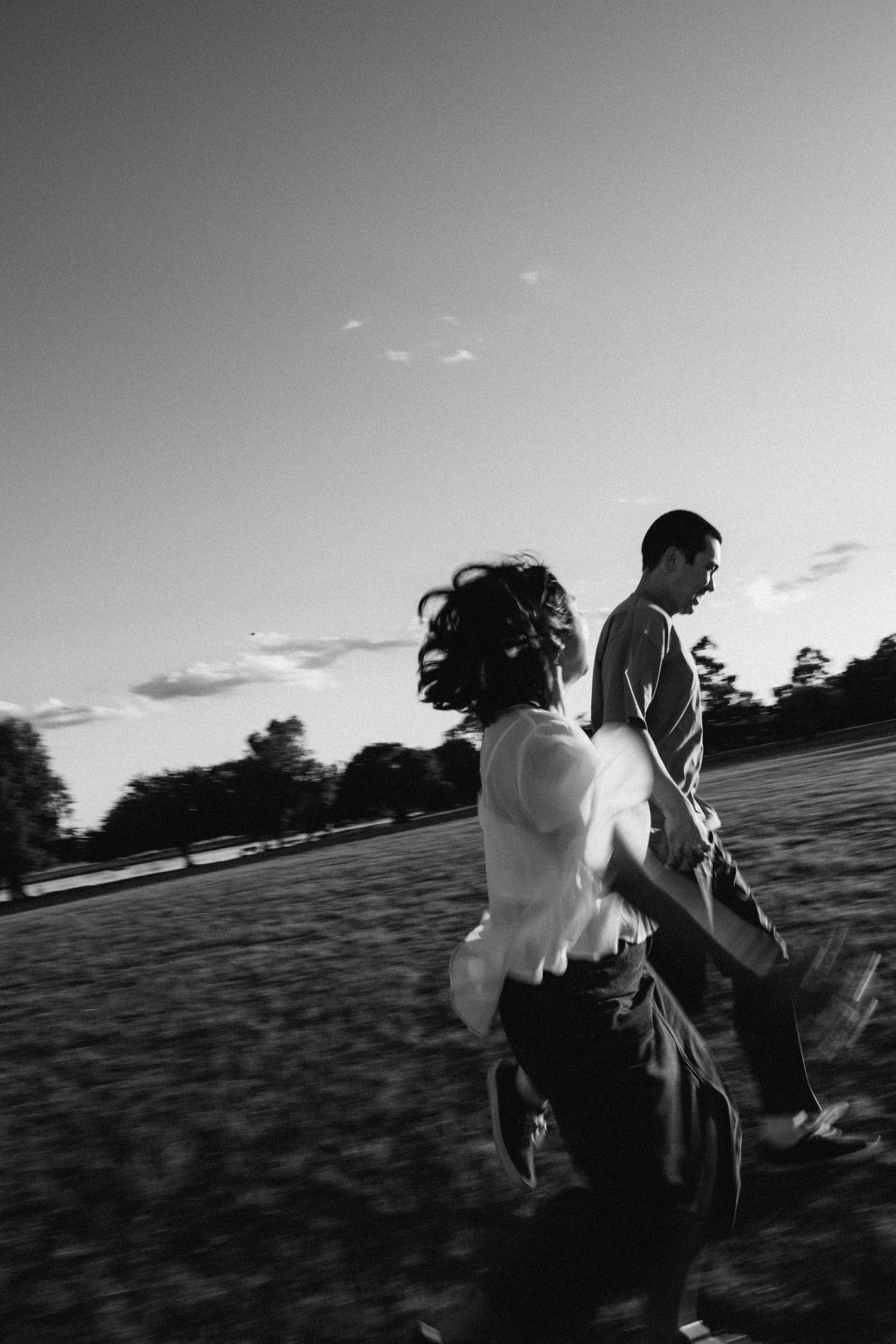Black and white photo of two people running outdoors, with a clear sky and trees in the background.
