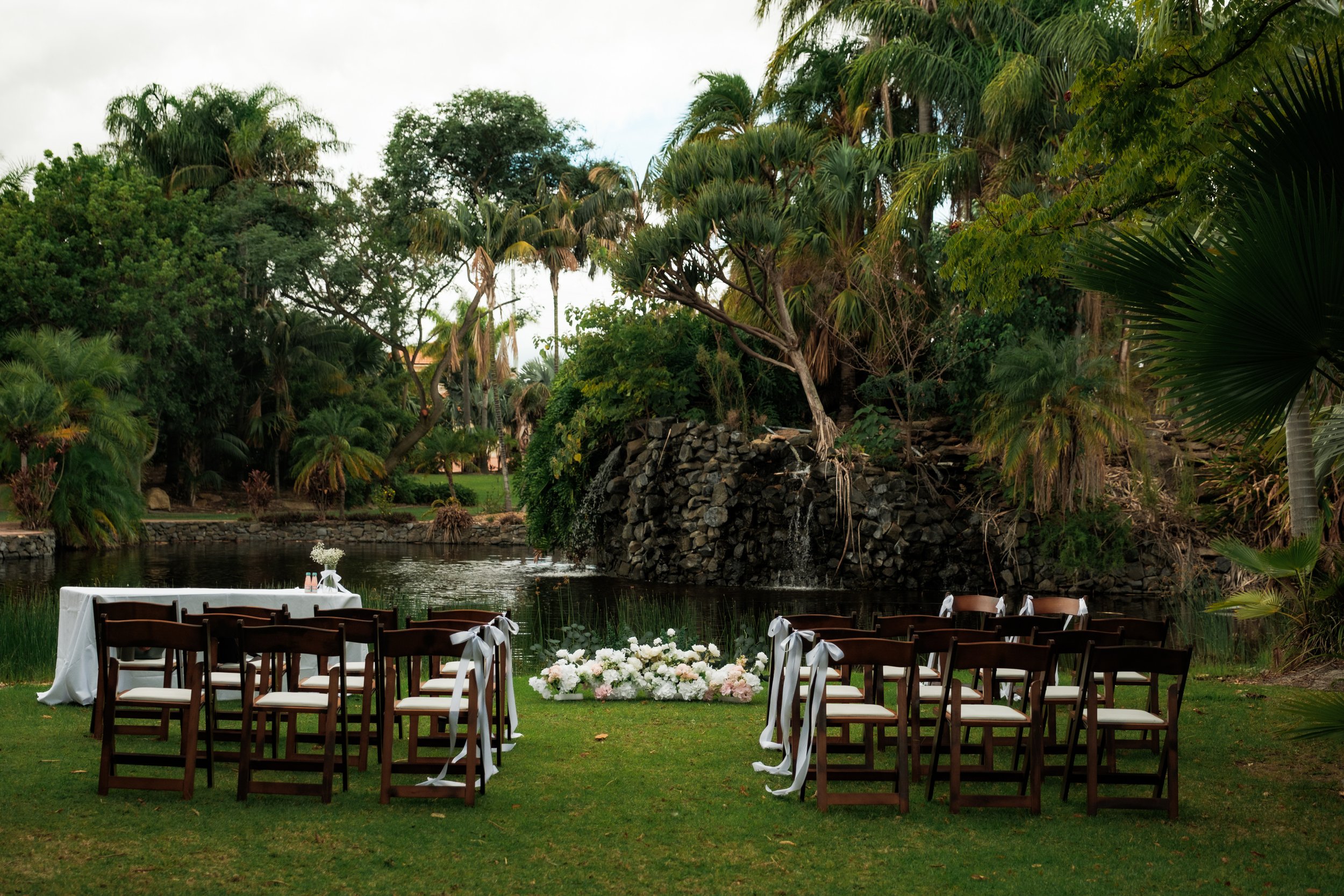 Wedding ceremony setup outdoors by a pond with chairs decorated with white ribbons, a floral arrangement, and surrounded by lush green trees and plants.