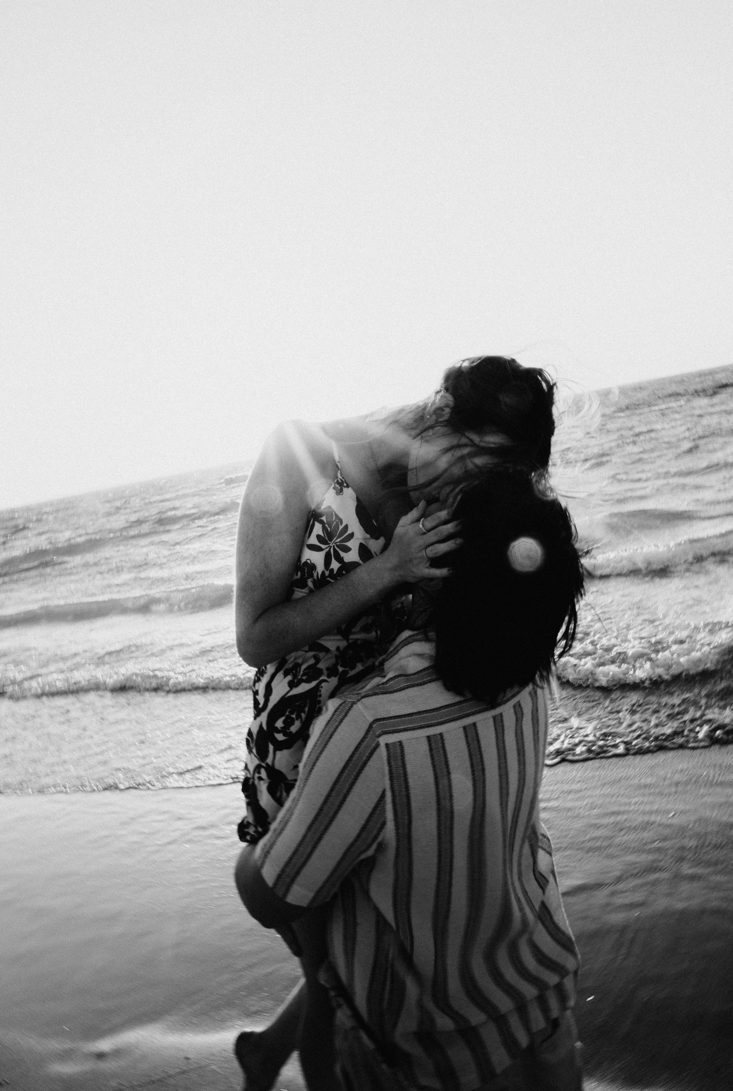 A woman and a young girl embracing on the beach near the ocean, with ocean waves visible in the background.