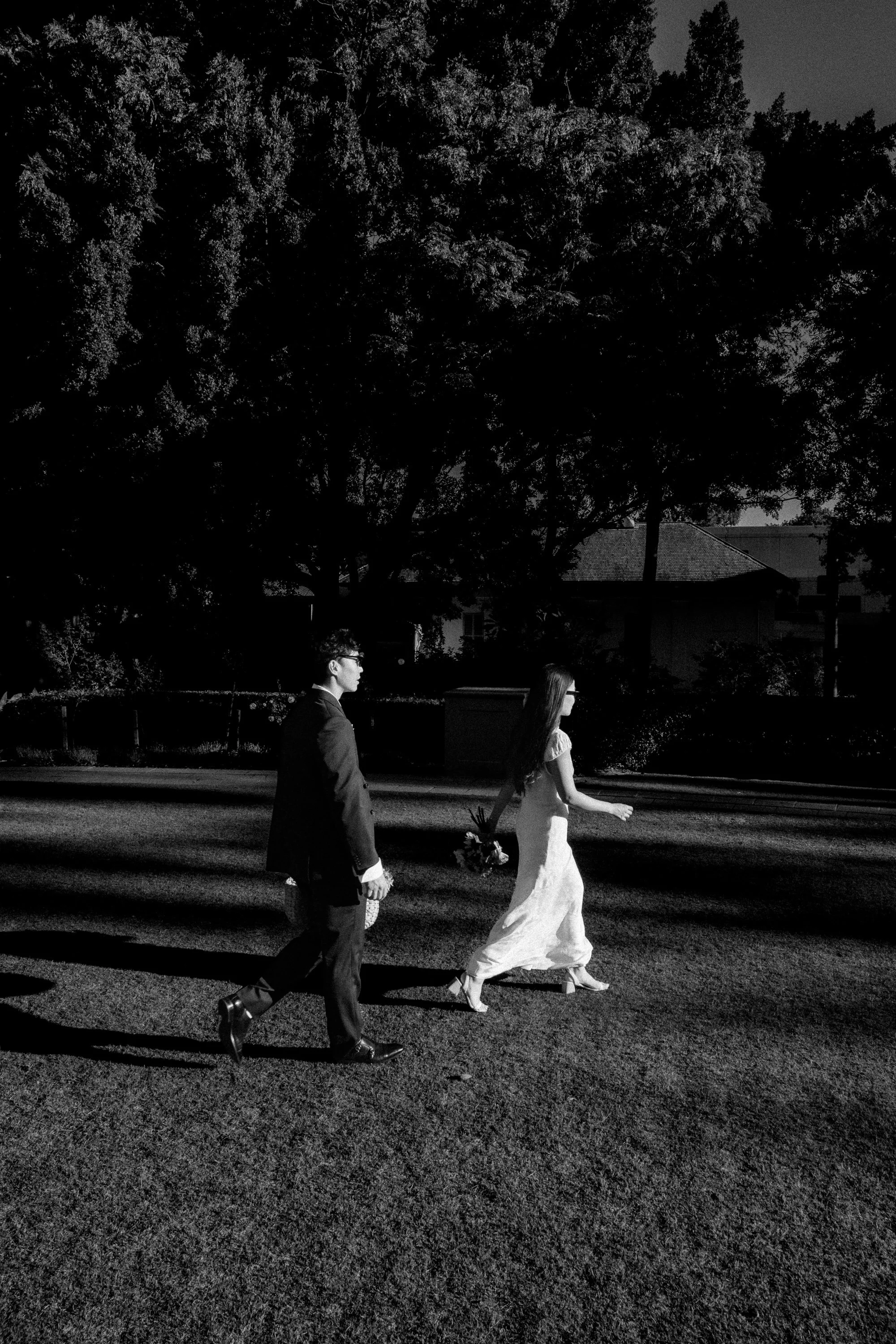 Black and white photo of a man and a woman walking across a street, holding hands, with trees and a house in the background.