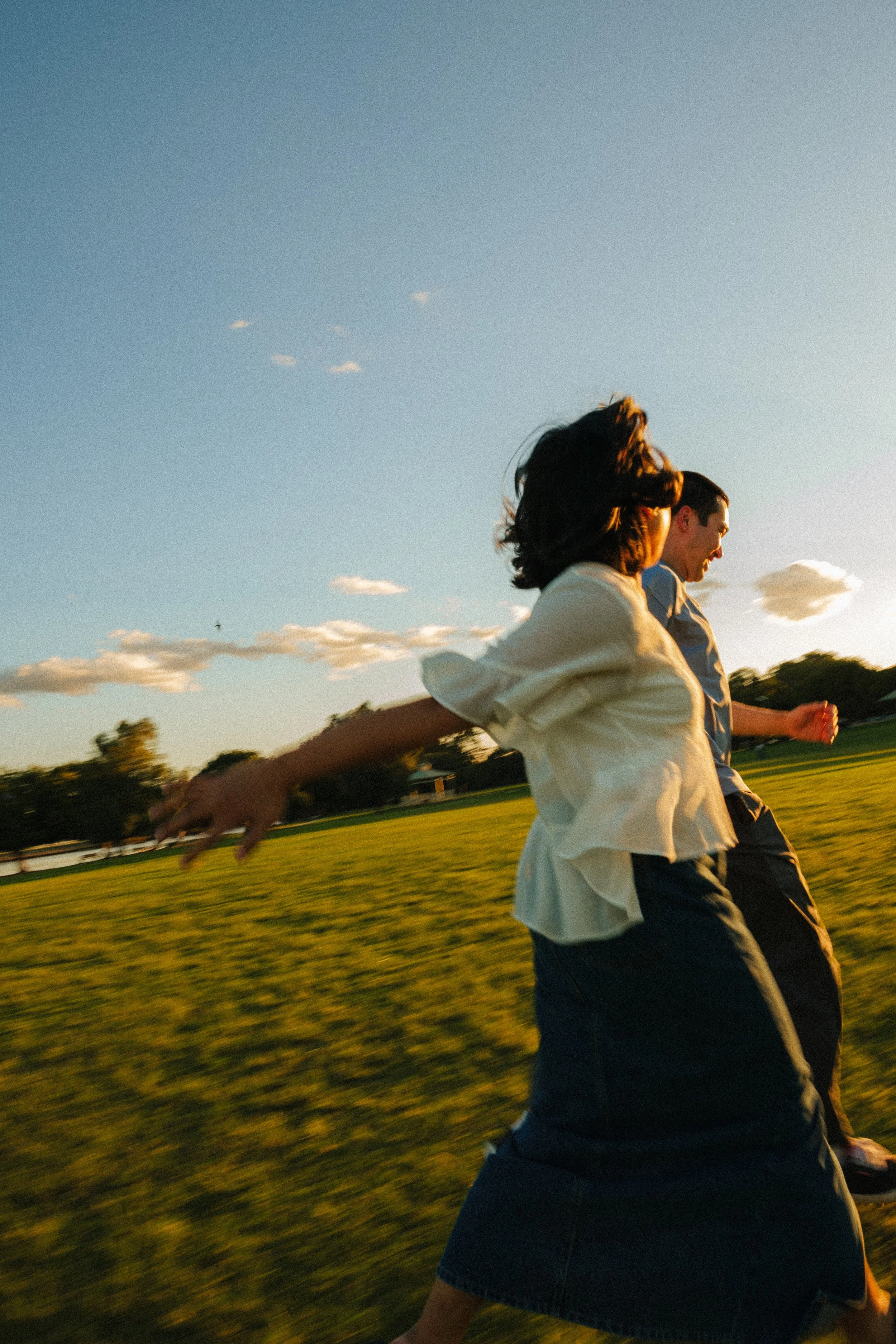 Two people, a woman and a man, are running outdoors on a grassy field during sunset or late afternoon. The woman has dark hair, is wearing a white shirt, and is in front. The man has short dark hair, is wearing a light blue shirt, and is behind her. Both appear happy and are enjoying the moment. The sky is mostly clear with a few small clouds.