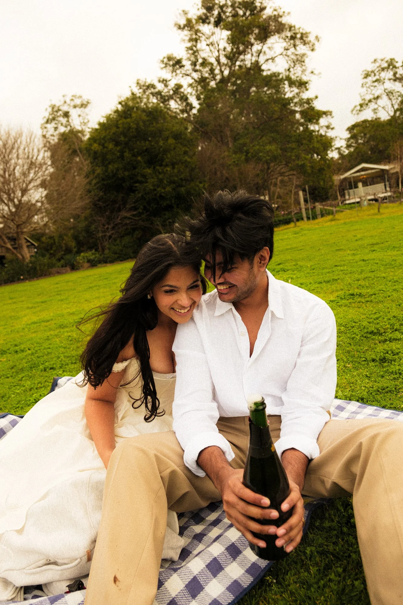 A young couple sitting on a checkered blanket in a grassy field, laughing and smiling with a champagne bottle, surrounded by trees and an overcast sky.