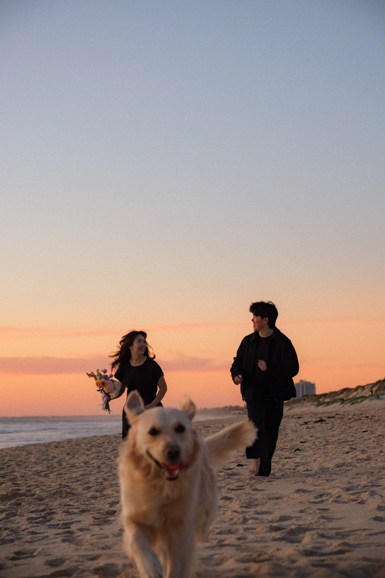 A couple and their dog running on a beach during sunset, the woman holding a bouquet of flowers, the man dressed in dark clothing, and the dog happily in front.