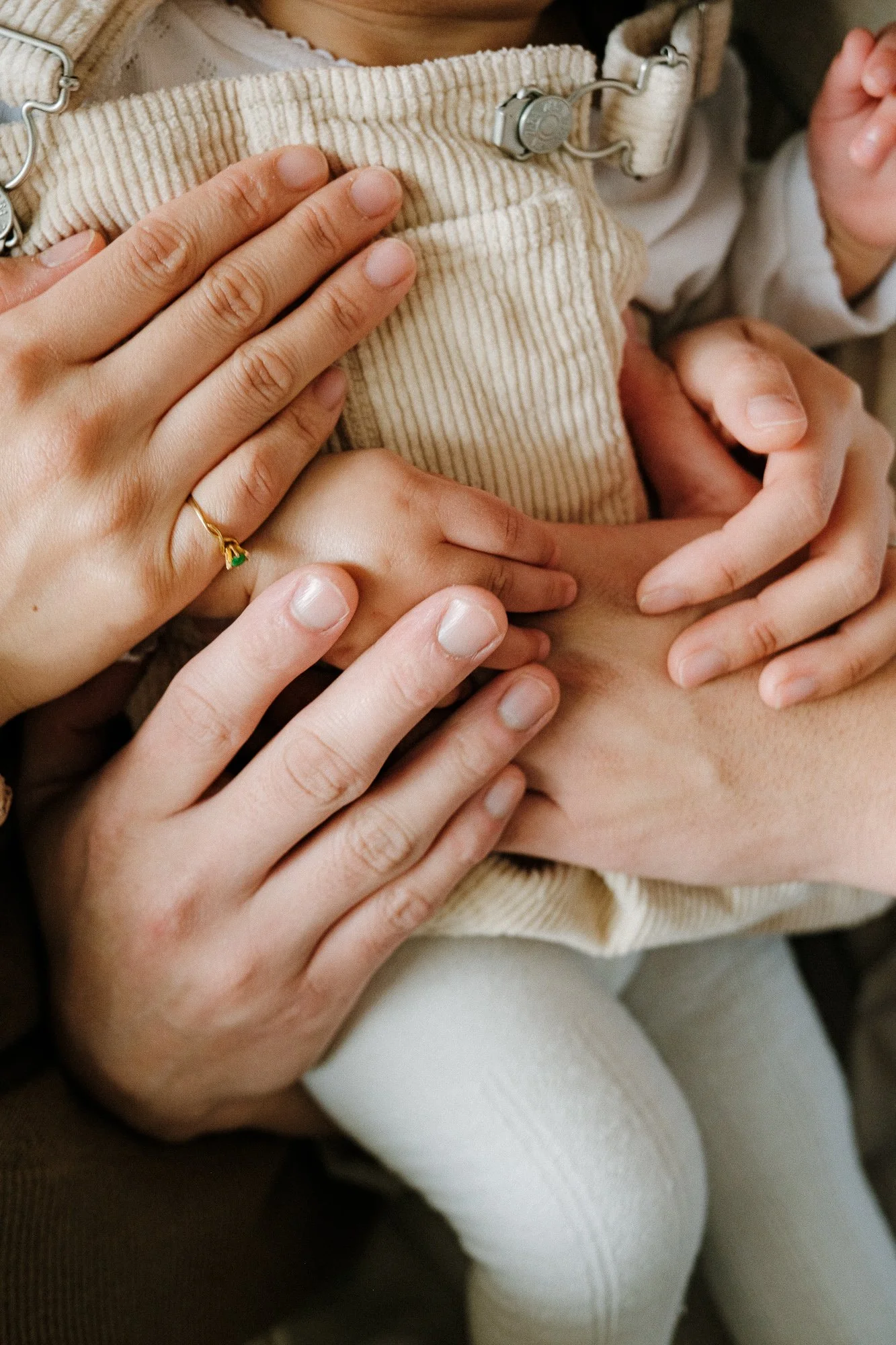 Close-up of multiple hands gently holding a child's midsection, with a focus on the hands, some wearing rings, on the child's tummy. The child is wearing a light-colored corduroy outfit.