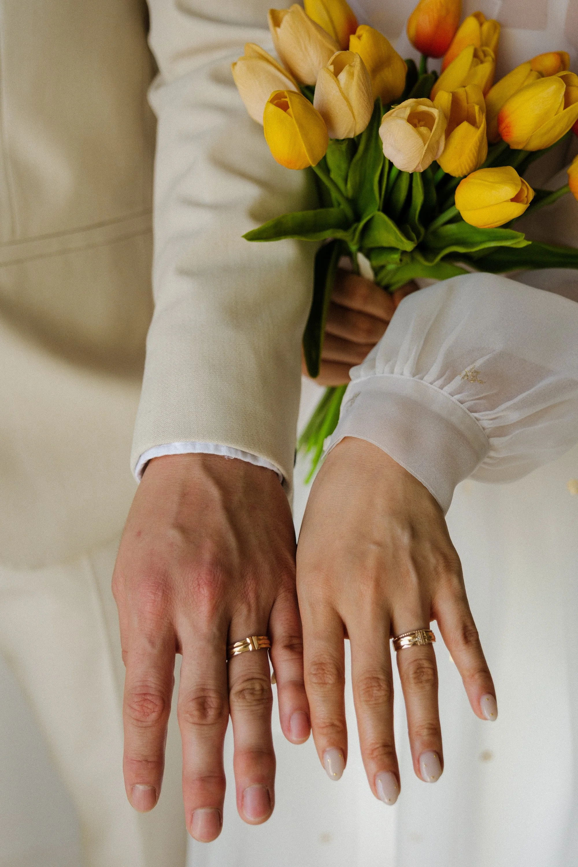 A person is holding a bouquet of yellow and cream tulips with their hands, wearing a white outfit with puffy sleeves and gold rings on their fingers.