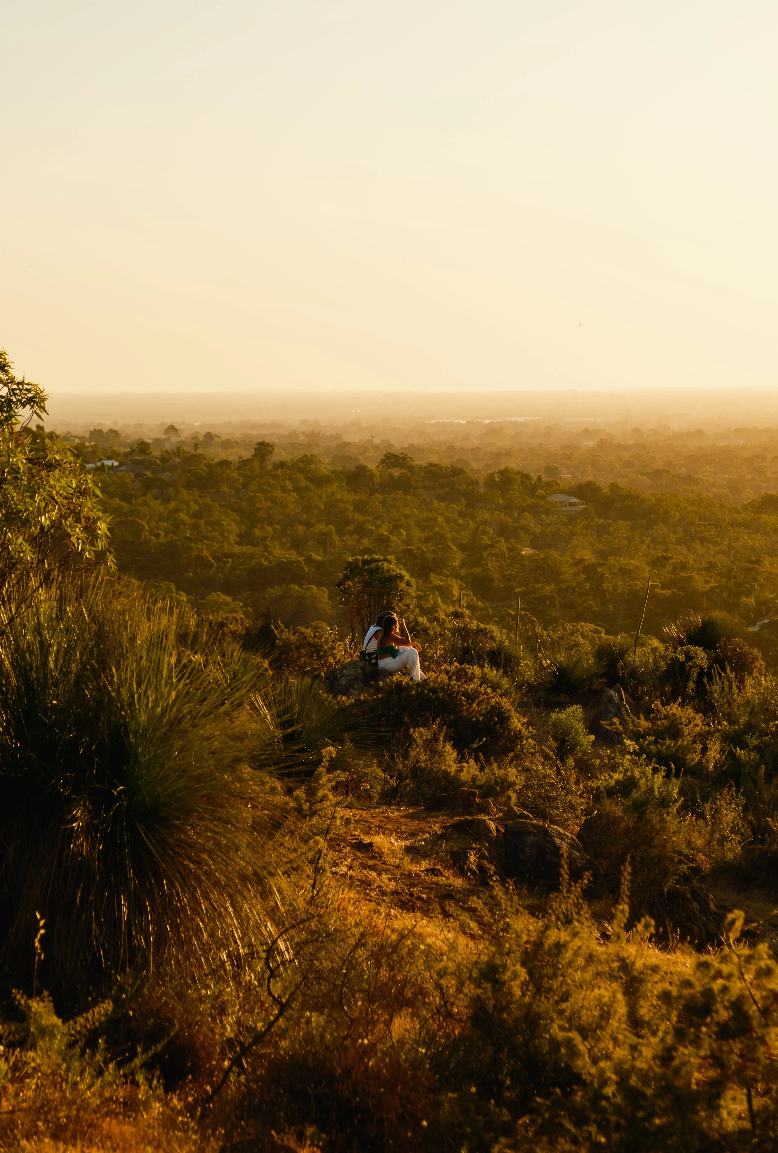 Two people sitting on a rocky hilltop overlooking a green, forested landscape at sunset.