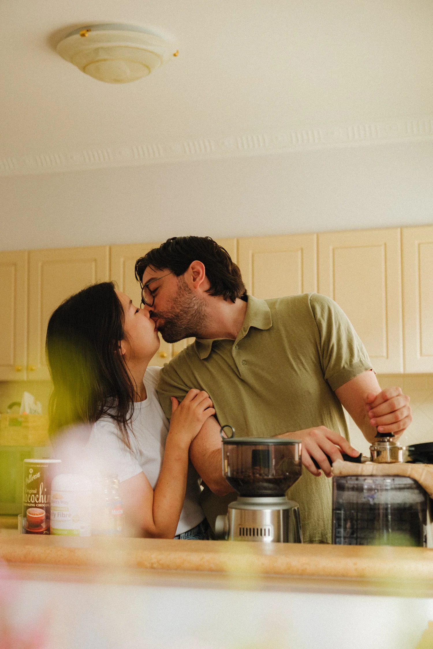 A couple sharing a kiss in a kitchen, with kitchen appliances, jars, and a woman wearing glasses.