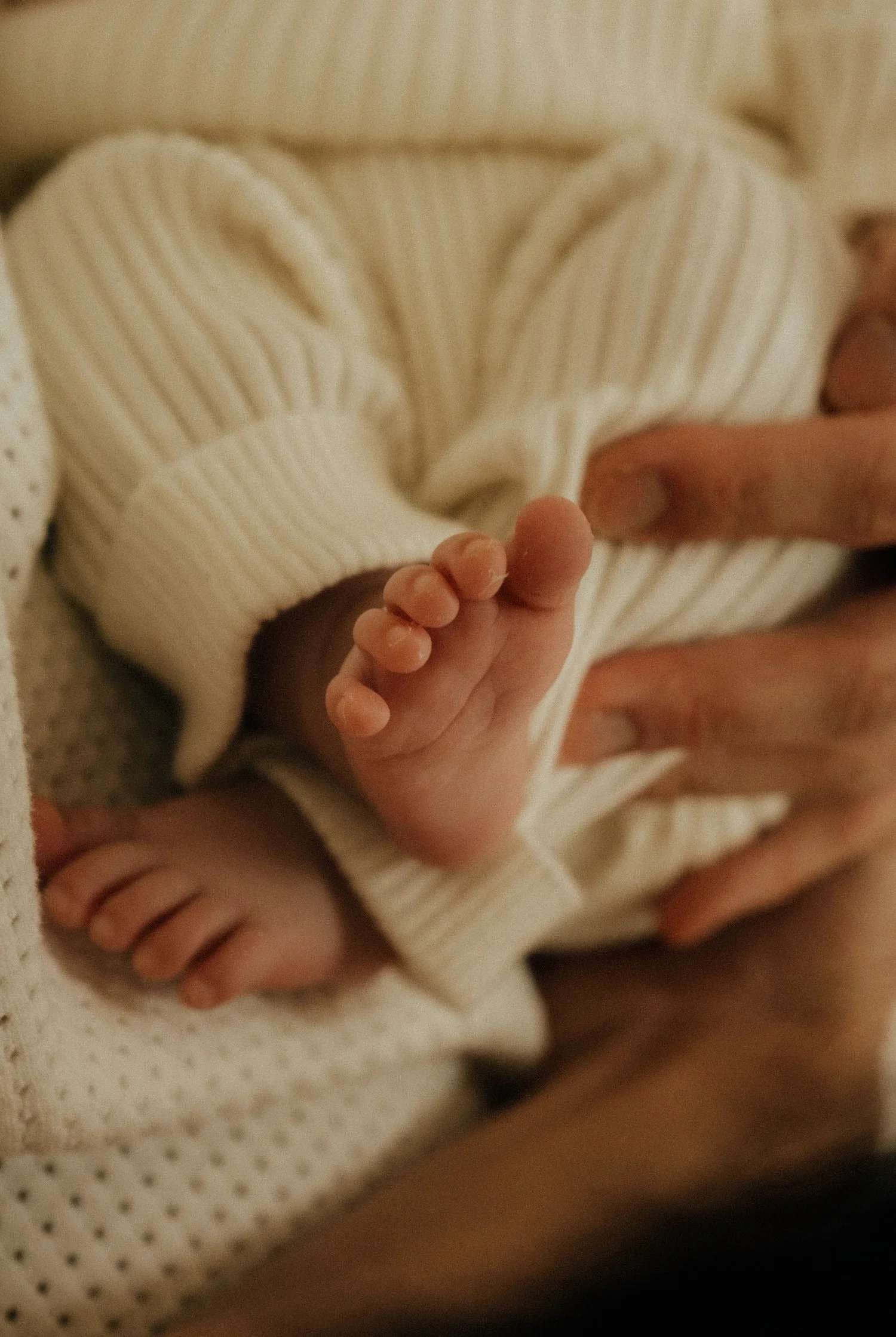 Close-up of a baby's feet being gently held by an adult's hand, with the baby wearing a cream-colored, ribbed, long-sleeve outfit.