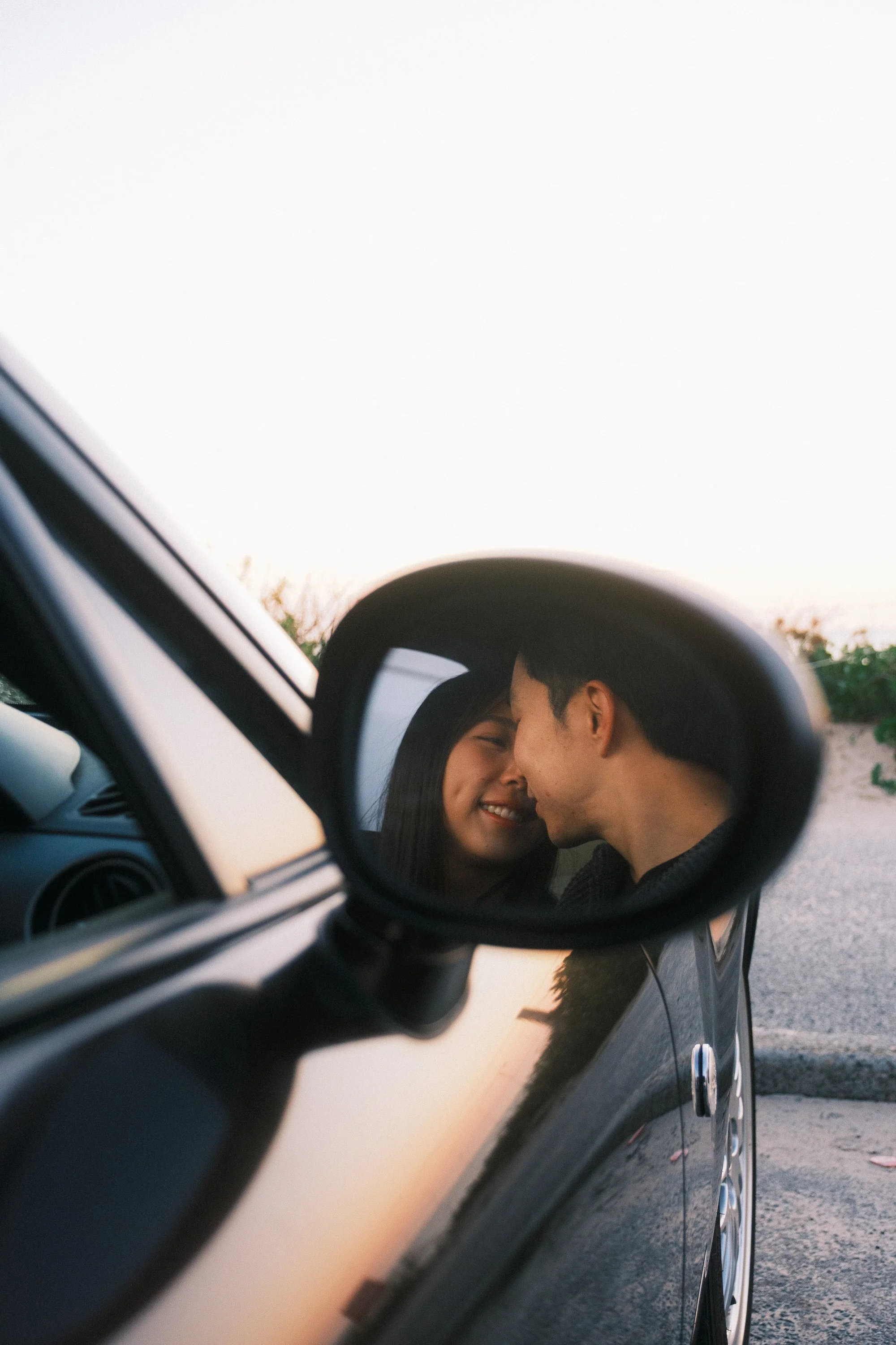 A couple is smiling and leaning close to each other inside a car, their reflection visible in the side mirror.