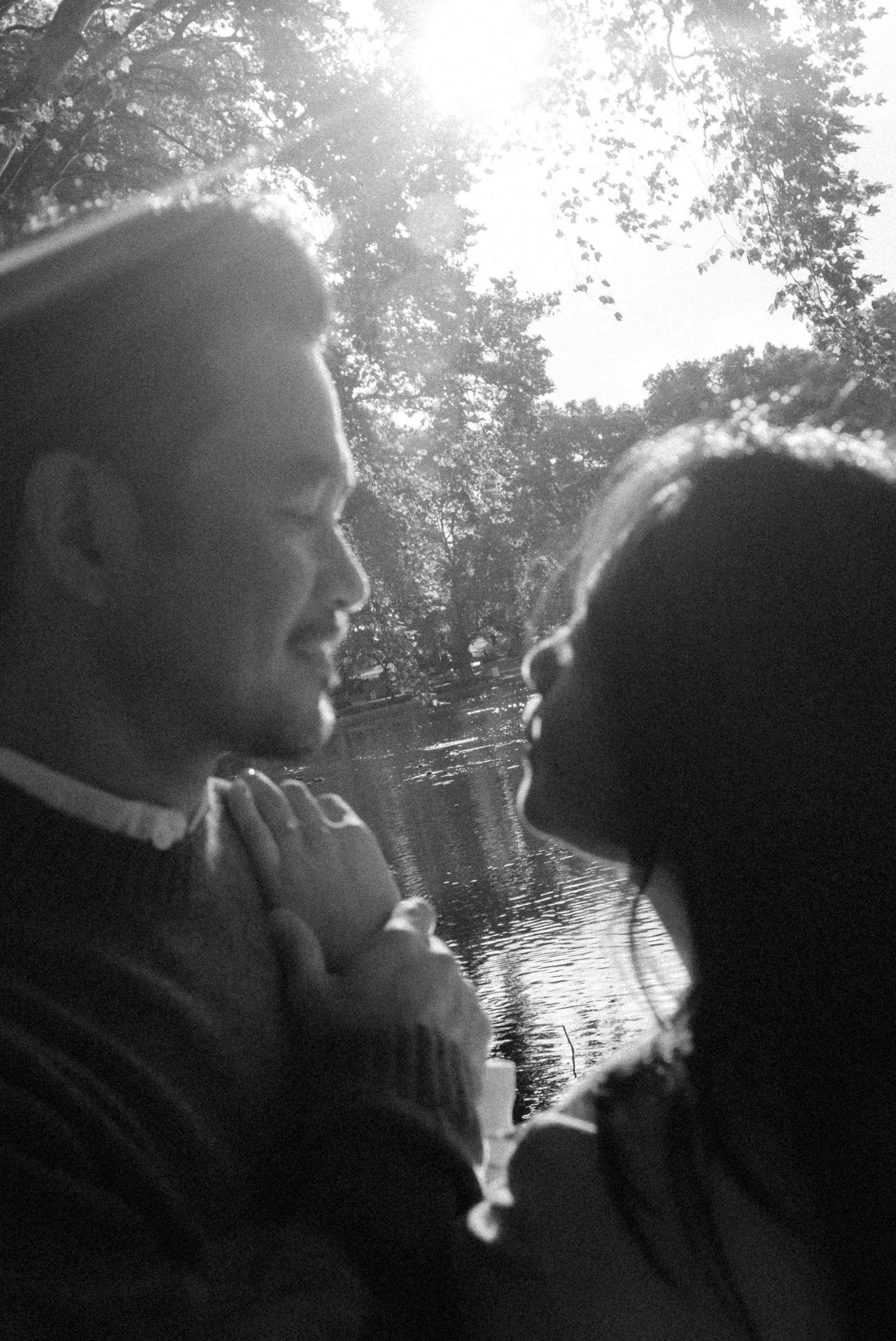Two children facing each other close together outdoors near a body of water, with trees and sunlight in the background, in black and white.