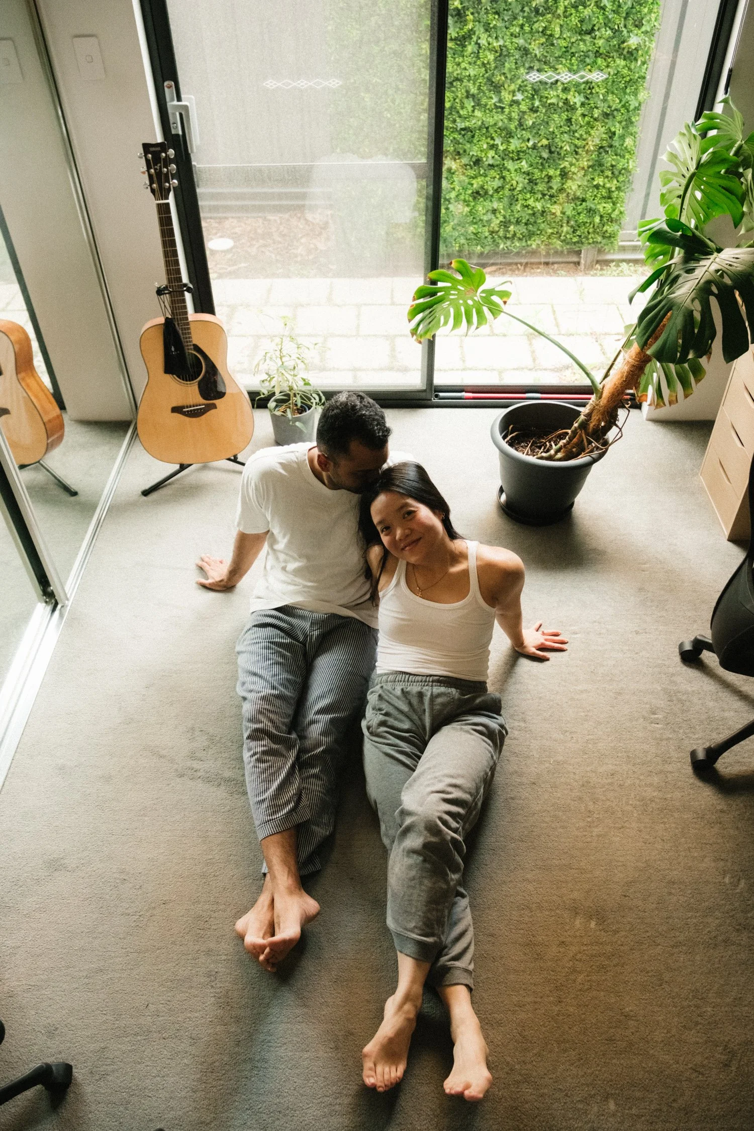 A man and woman sitting on the carpeted floor inside a room, looking at each other and smiling. The man is leaning on his left arm, wearing a white t-shirt and striped pants. The woman is leaning back with her arms supporting her, wearing a white tank top and gray pants. Behind them is a large window showing green foliage outside. There is a potted plant and two guitars in the room.