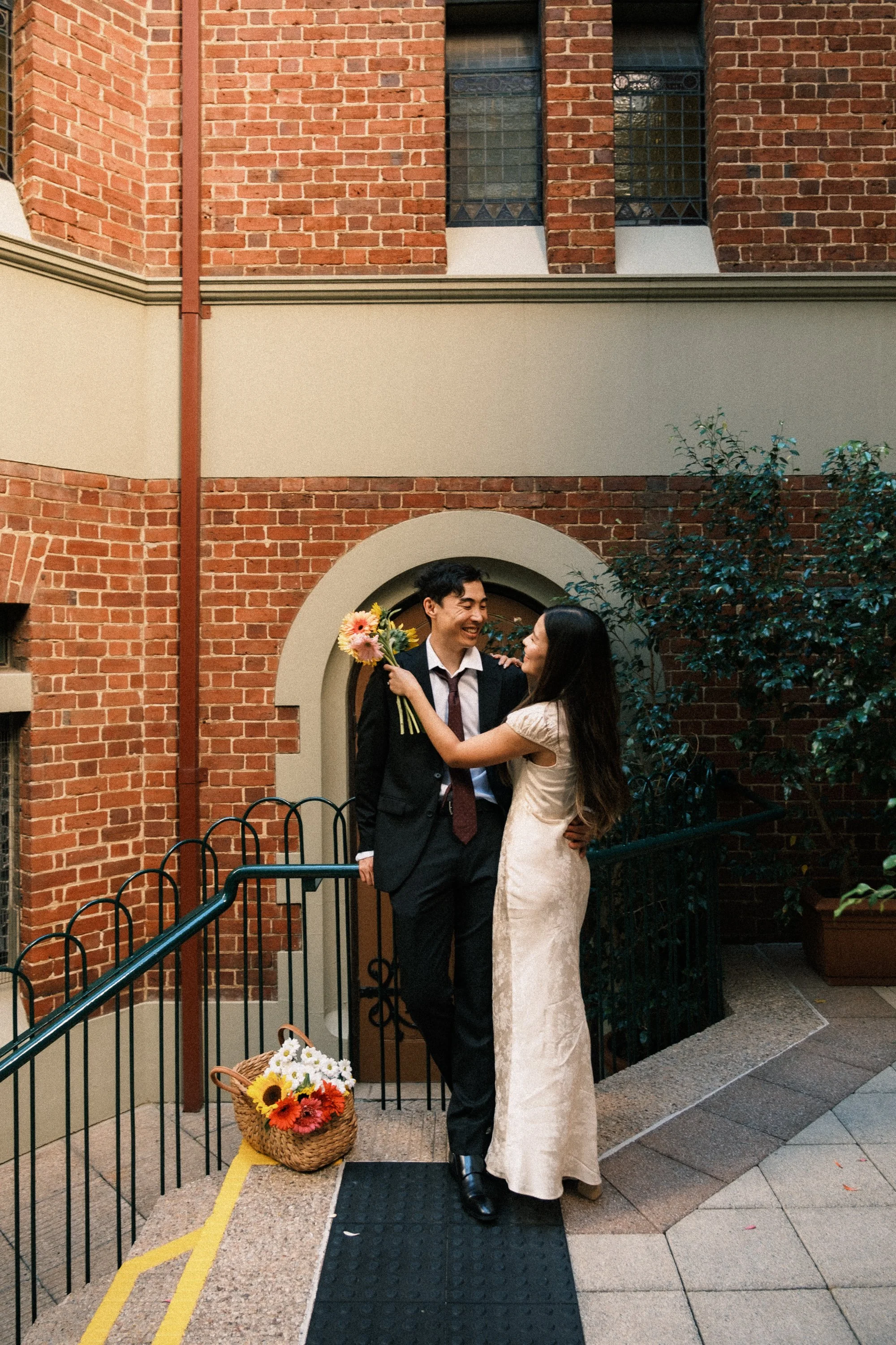 A joyful couple stands outside a brick building in a romantic embrace, with the woman holding a bouquet of flowers and a basket of colorful flowers on the ground nearby.