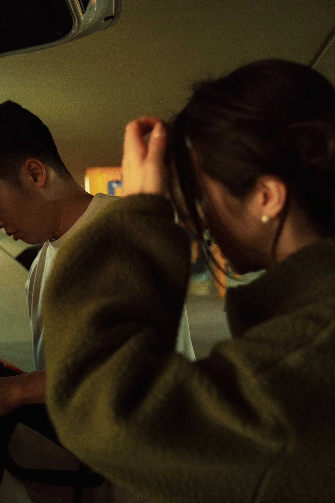 Two people sitting closely together, one with dark hair and an earring, looking downward, and the other with short hair, in a dimly lit indoor setting.