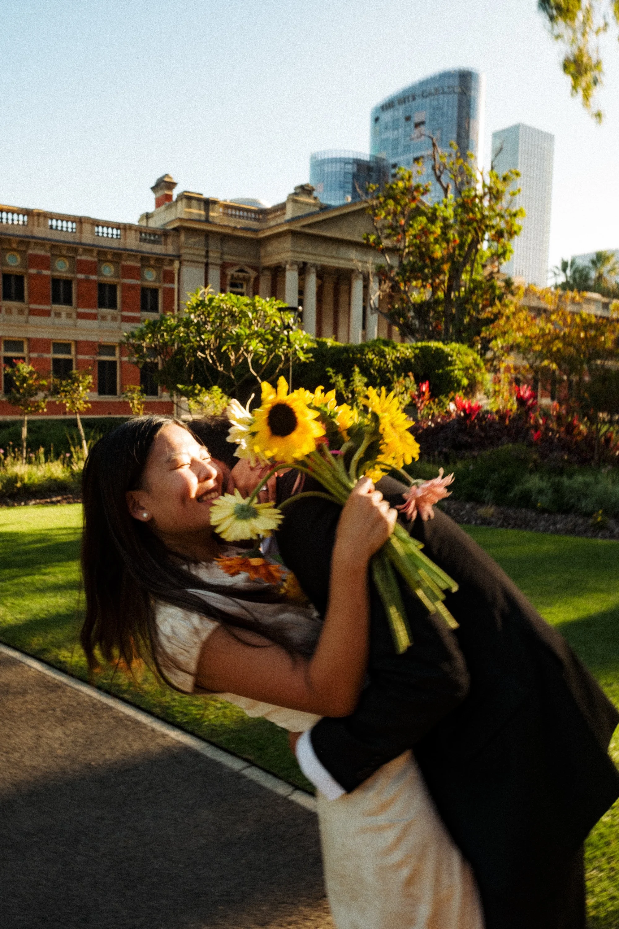 A woman with long dark hair smiling and holding a bouquet of yellow flowers while lying on green grass in a park with an ornate building and tall modern skyscrapers in the background.