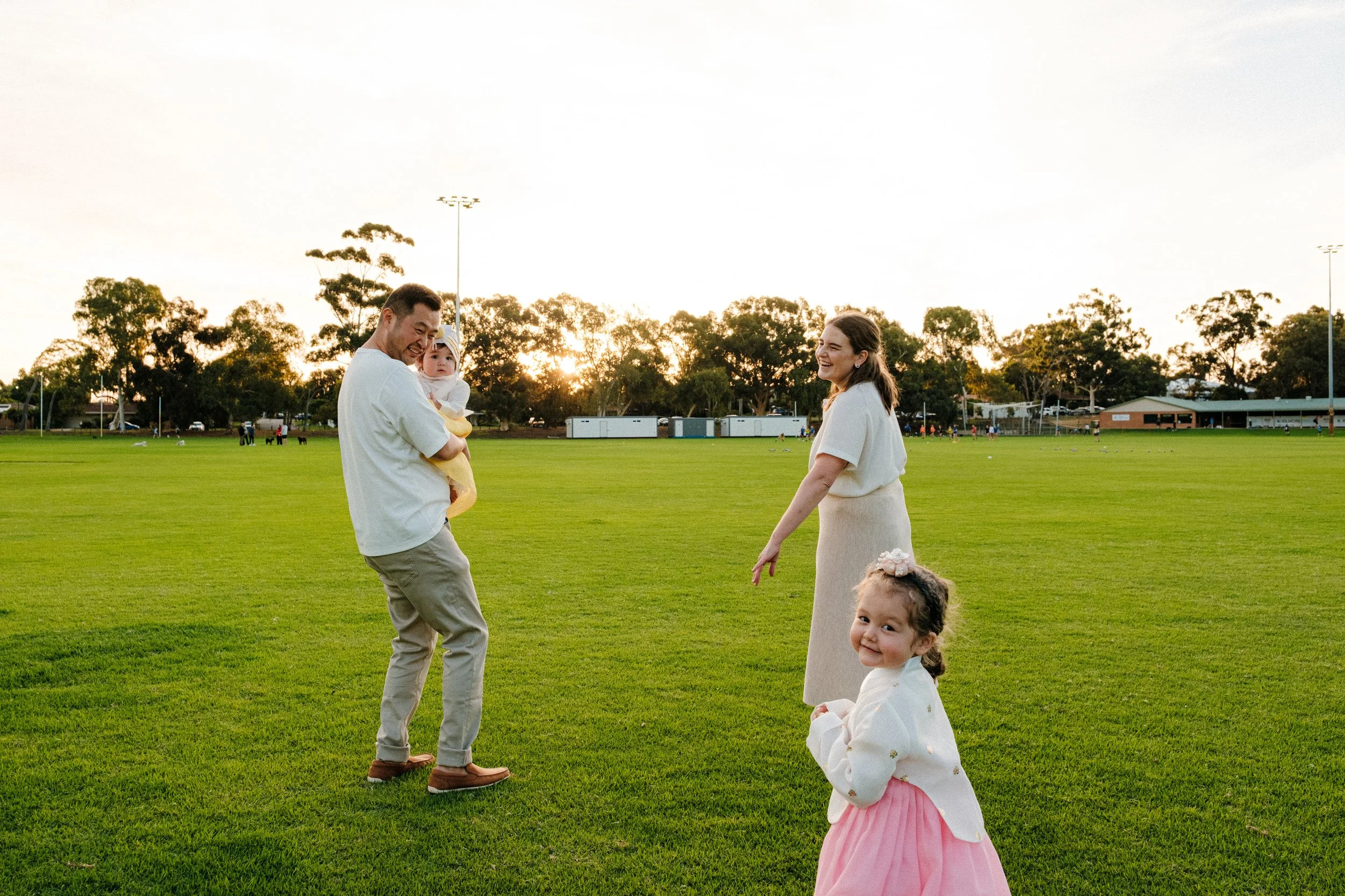A family enjoying time in a park during sunset, with a father holding a baby girl, a woman smiling, and a young girl in a pink skirt in the foreground.