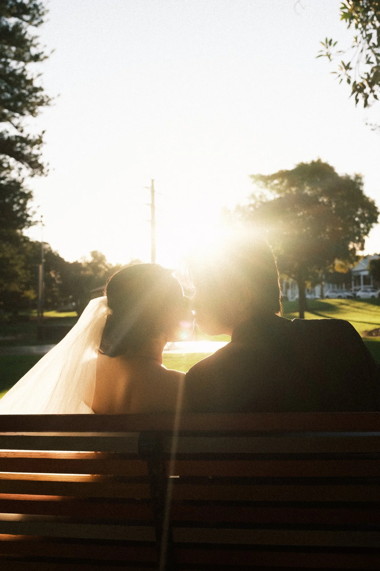 A couple sitting on a park bench, sharing a kiss at sunset. The bride is wearing a wedding veil, and the groom is in a dark suit. The sun is shining brightly behind them, creating a romantic glow.