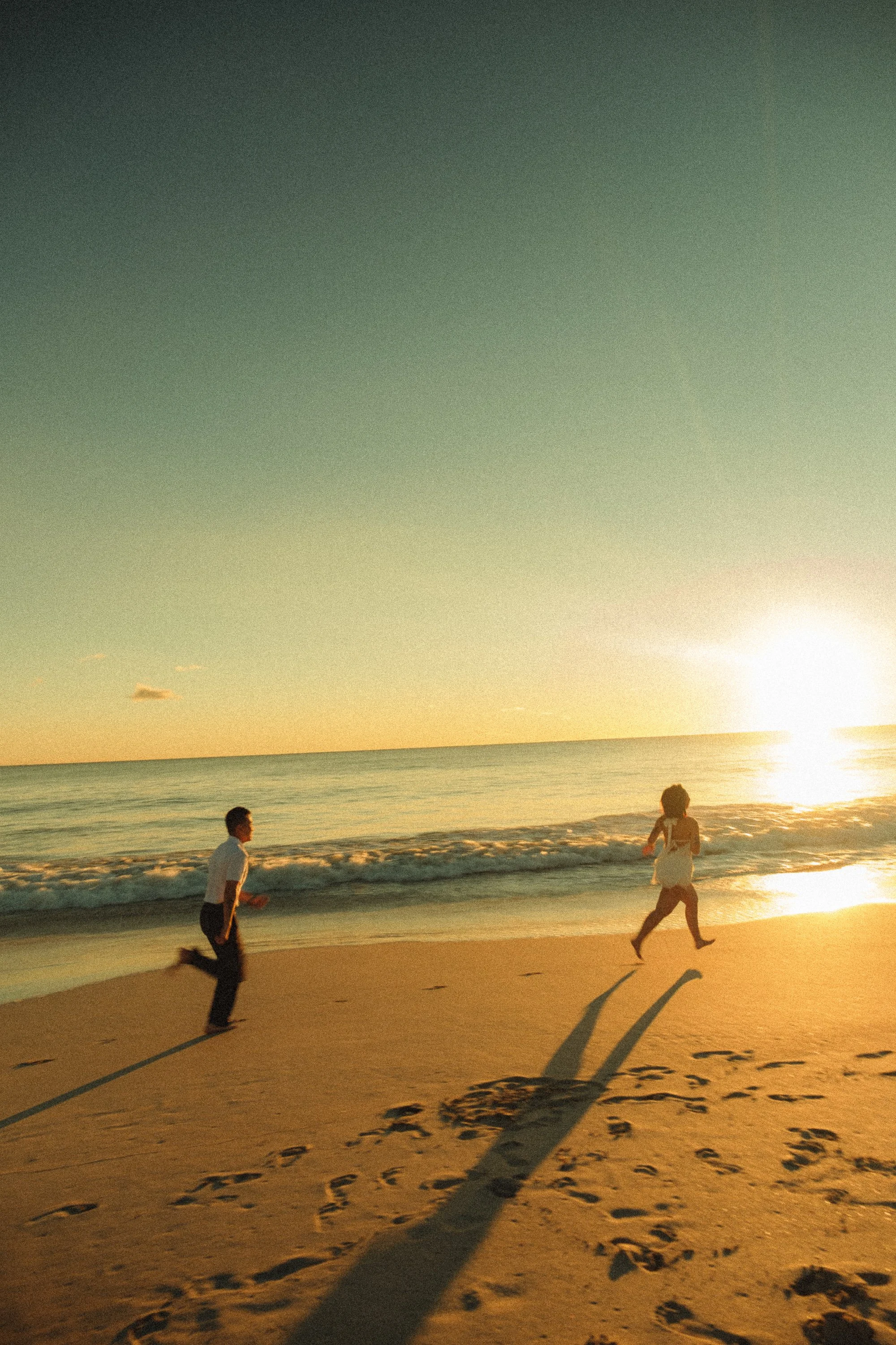 Children playing and running on the sandy beach during sunset with footprints and long shadows, ocean waves in the background.