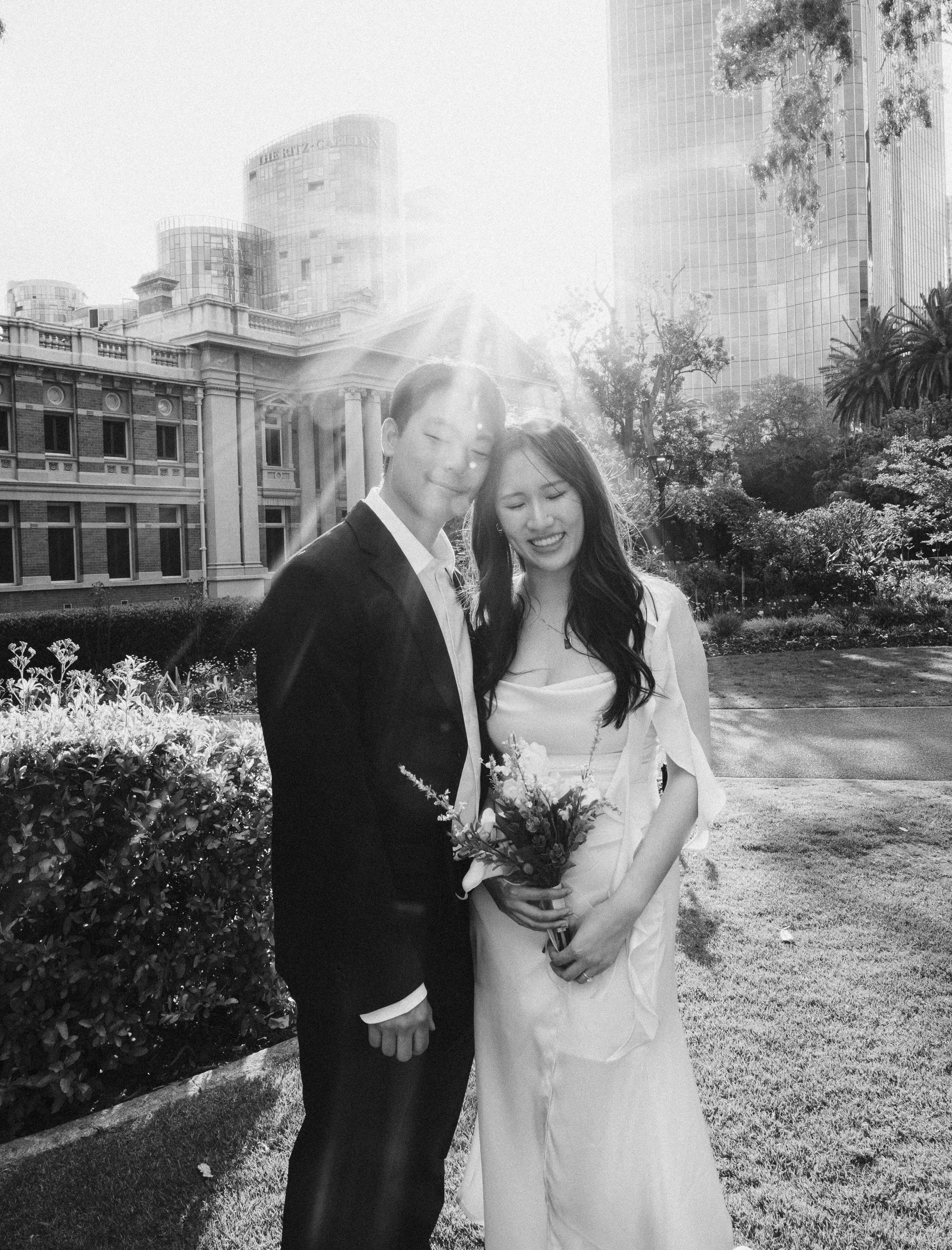 A black-and-white photo of a couple on their wedding day outdoors, with the city skyline in the background. The groom is wearing a suit, and the bride is in a white dress holding a bouquet of flowers, both smiling with their eyes closed as sunlight shines behind them.