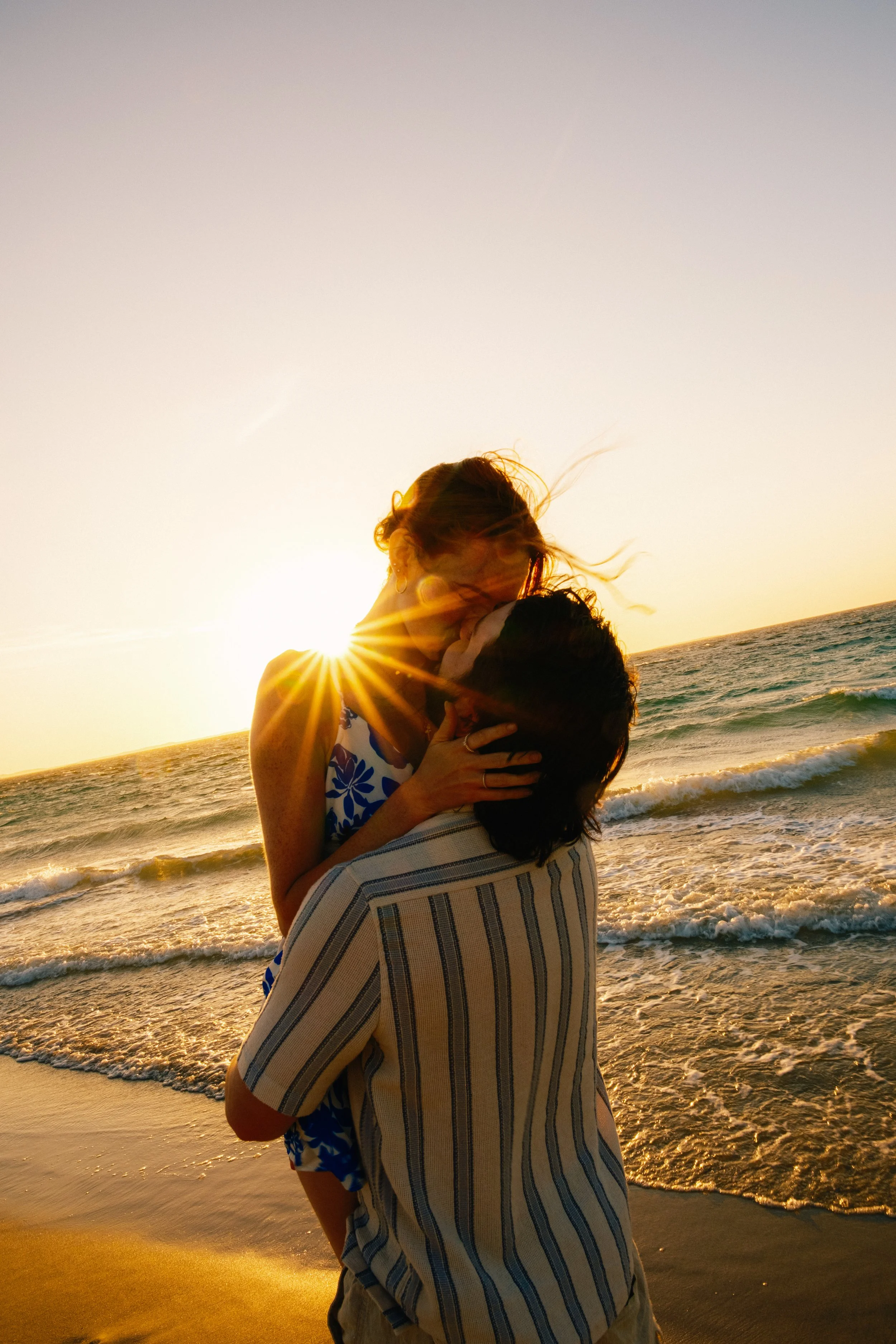 A couple is embracing and kissing on the beach at sunset, with the ocean waves in the background.