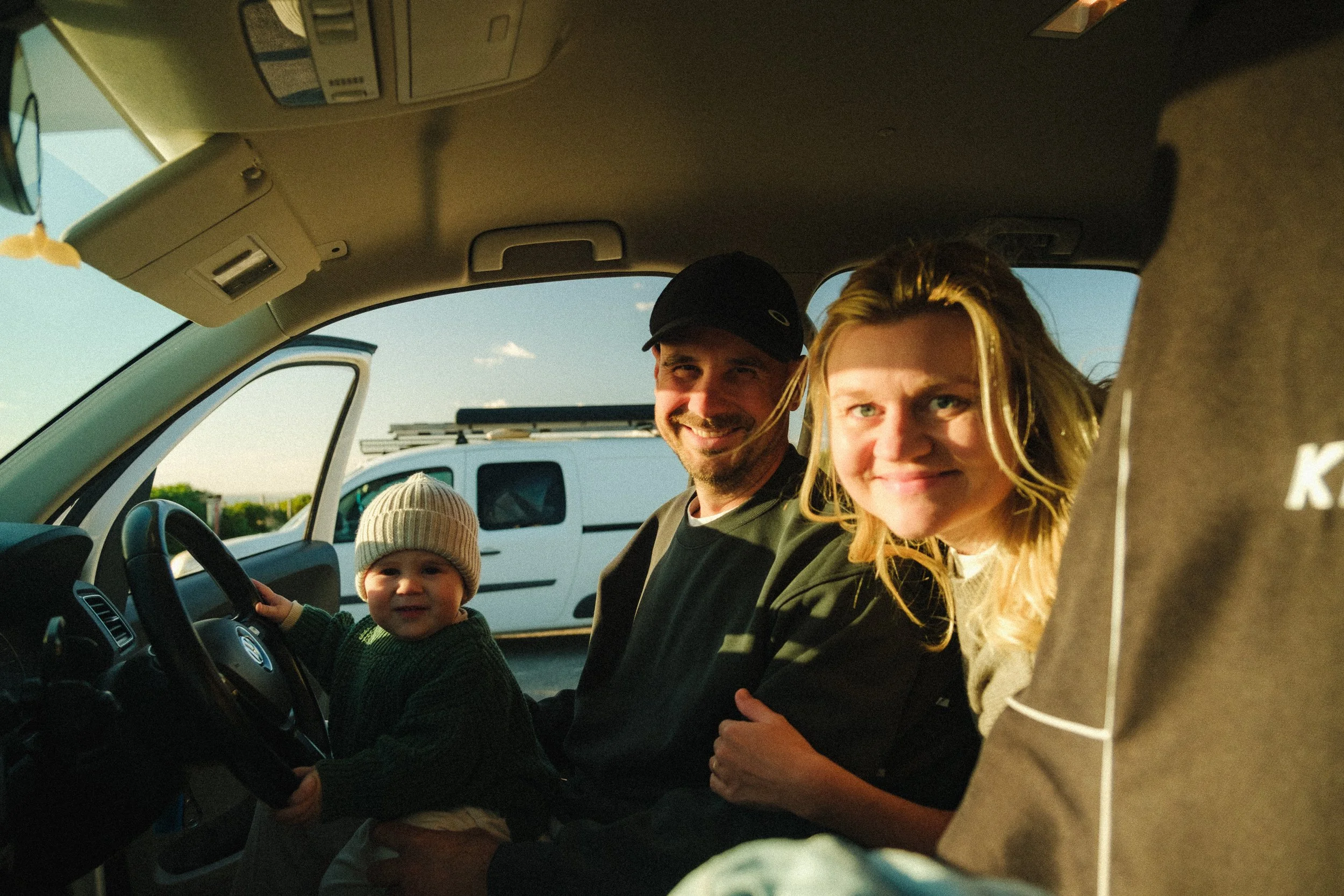 A family inside a vehicle, including a young boy in a driver’s seat, a woman with blonde hair, a man in a black cap, and part of another child, smiling and looking at the camera on a sunny day.