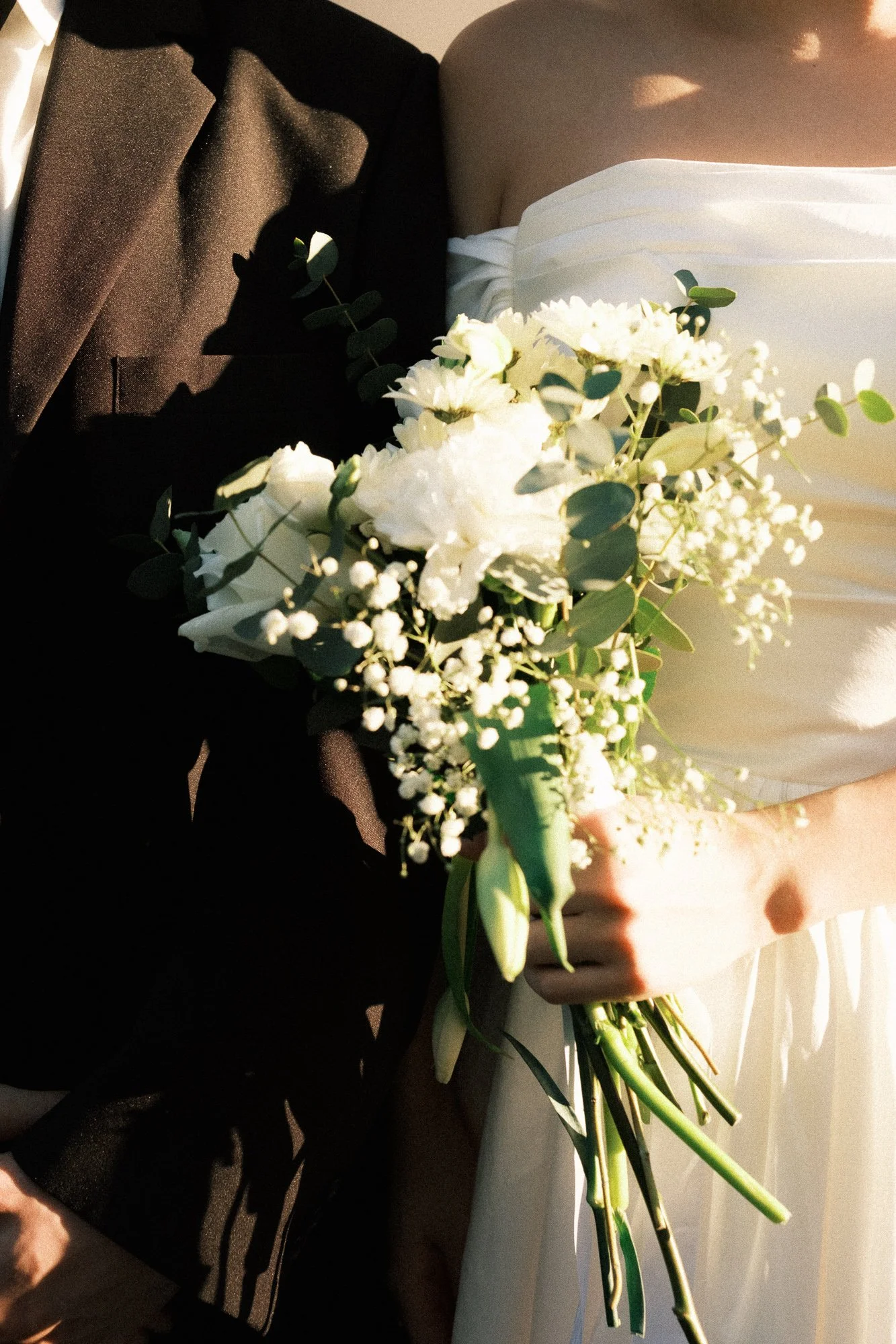 A bride holding a bouquet of white flowers, including lilies and baby's breath, with a shadow on her black jacket and sunlight on her white dress.