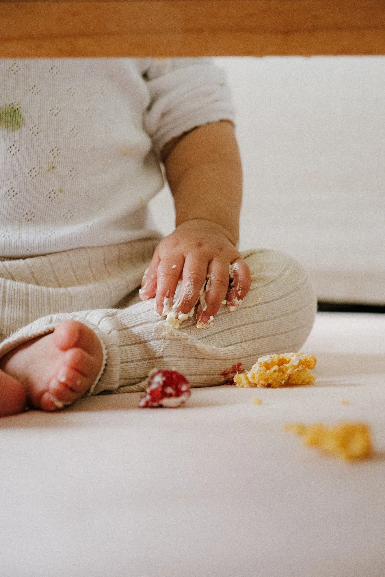 Close-up of a child's hand and foot, sitting on a light-colored surface, playing with cake crumbs and frosting, with part of their white shirt and beige striped pants visible.