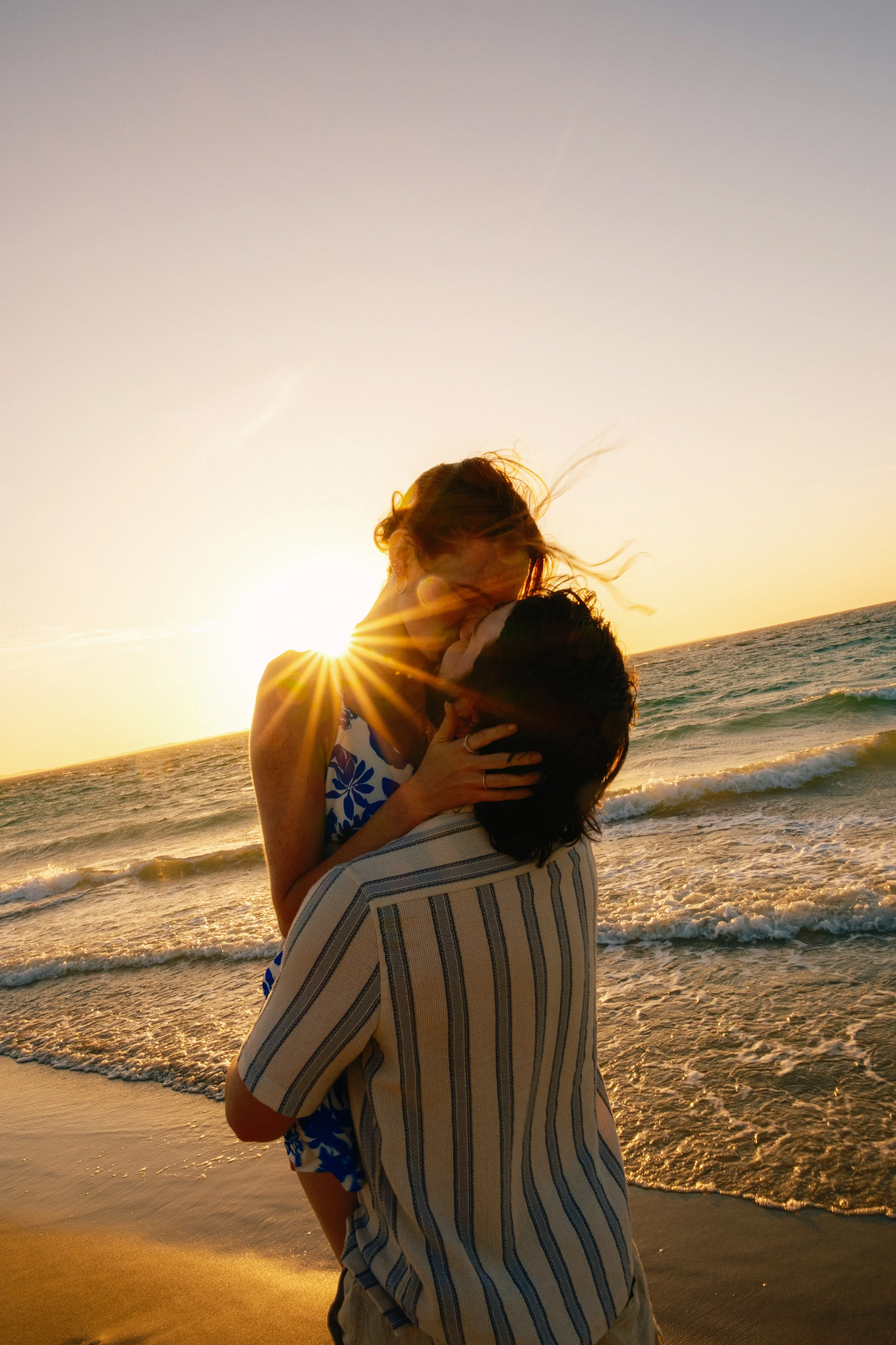 A couple embracing and kissing at the beach during sunset, with the sun shining behind them and gentle waves in the background.