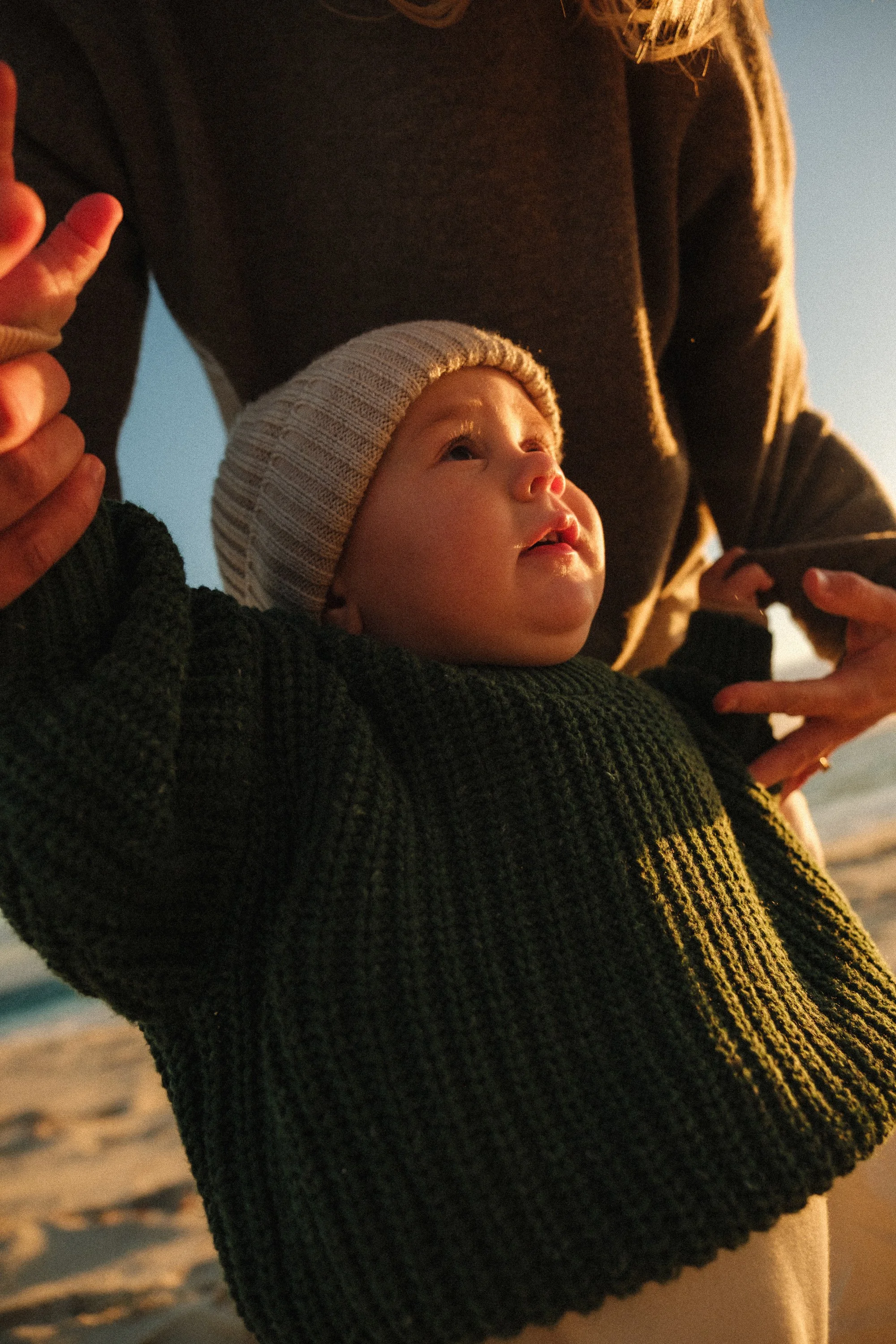 A close-up of a baby with fair skin, wearing a striped knit hat and a dark green chunky sweater, looking up with a curious expression near sunset with a blurred beach background.