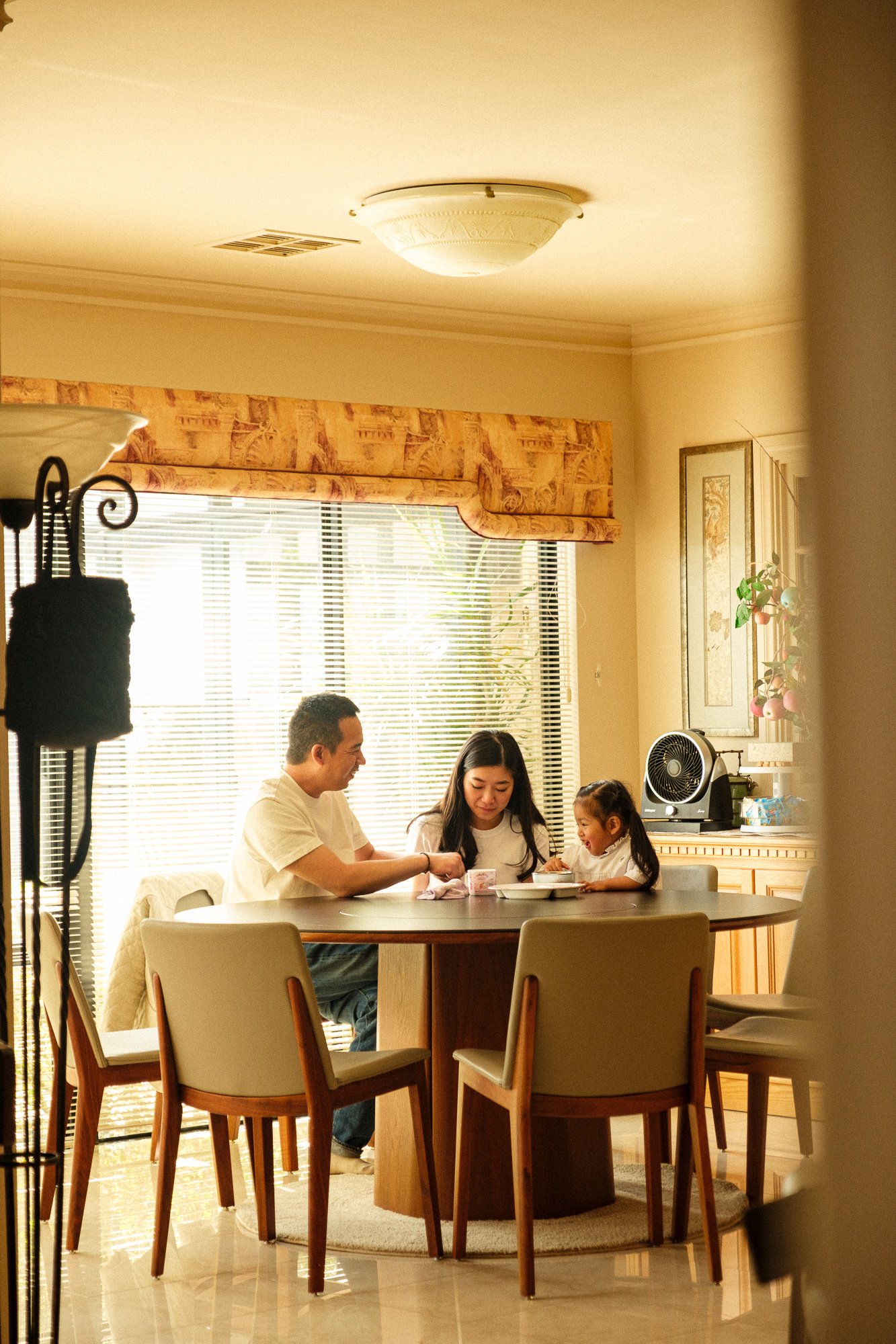 Family of four sitting at a dining table, sharing a meal, in a bright, cozy kitchen with window blinds, decorative curtains, and a cabinet with ornaments.