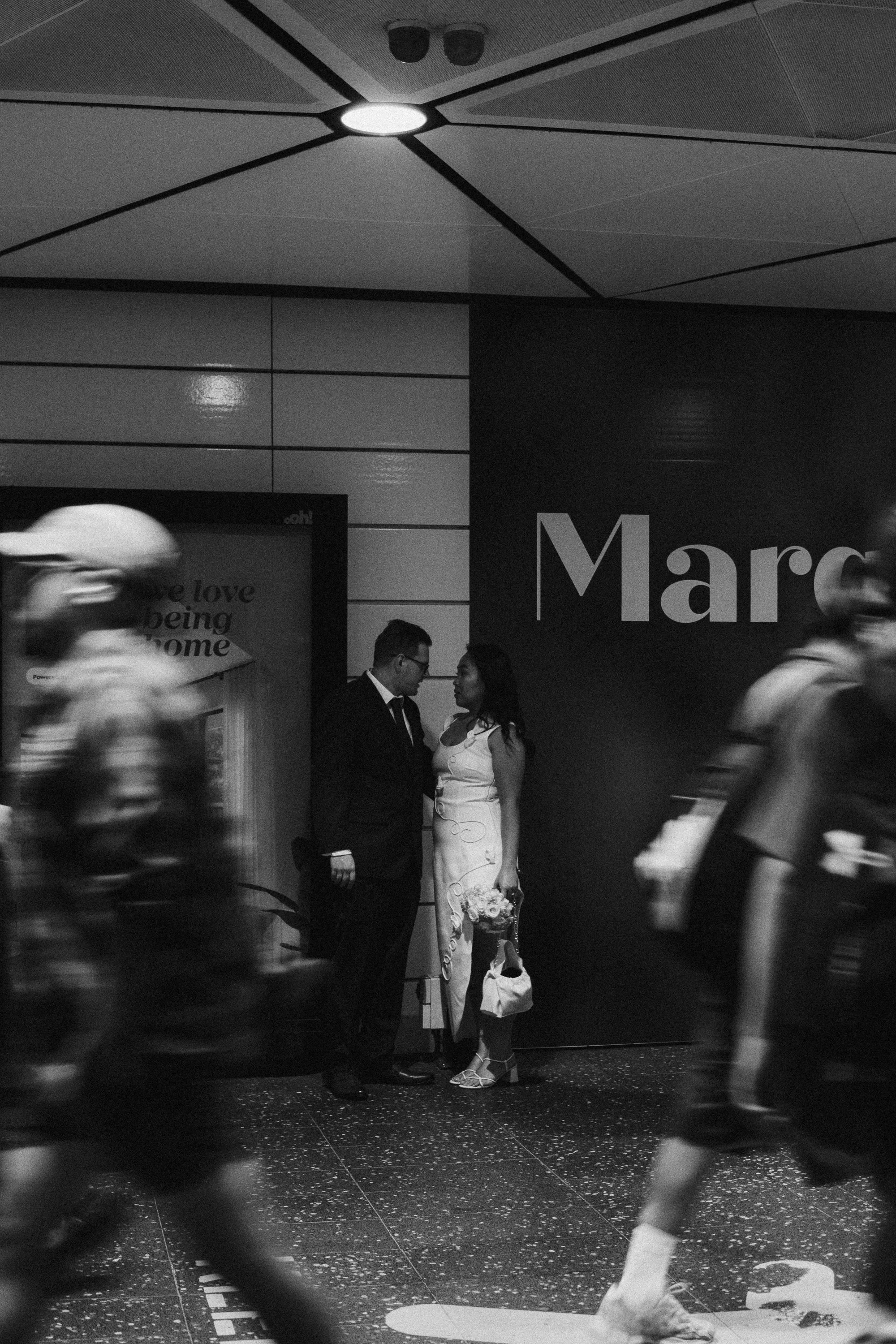 A couple dressed in wedding attire stands close together, facing each other, in a busy urban setting with blurred pedestrians walking past. The man is wearing a suit and glasses, holding a cup, while the woman is in a white dress holding a bouquet of flowers.