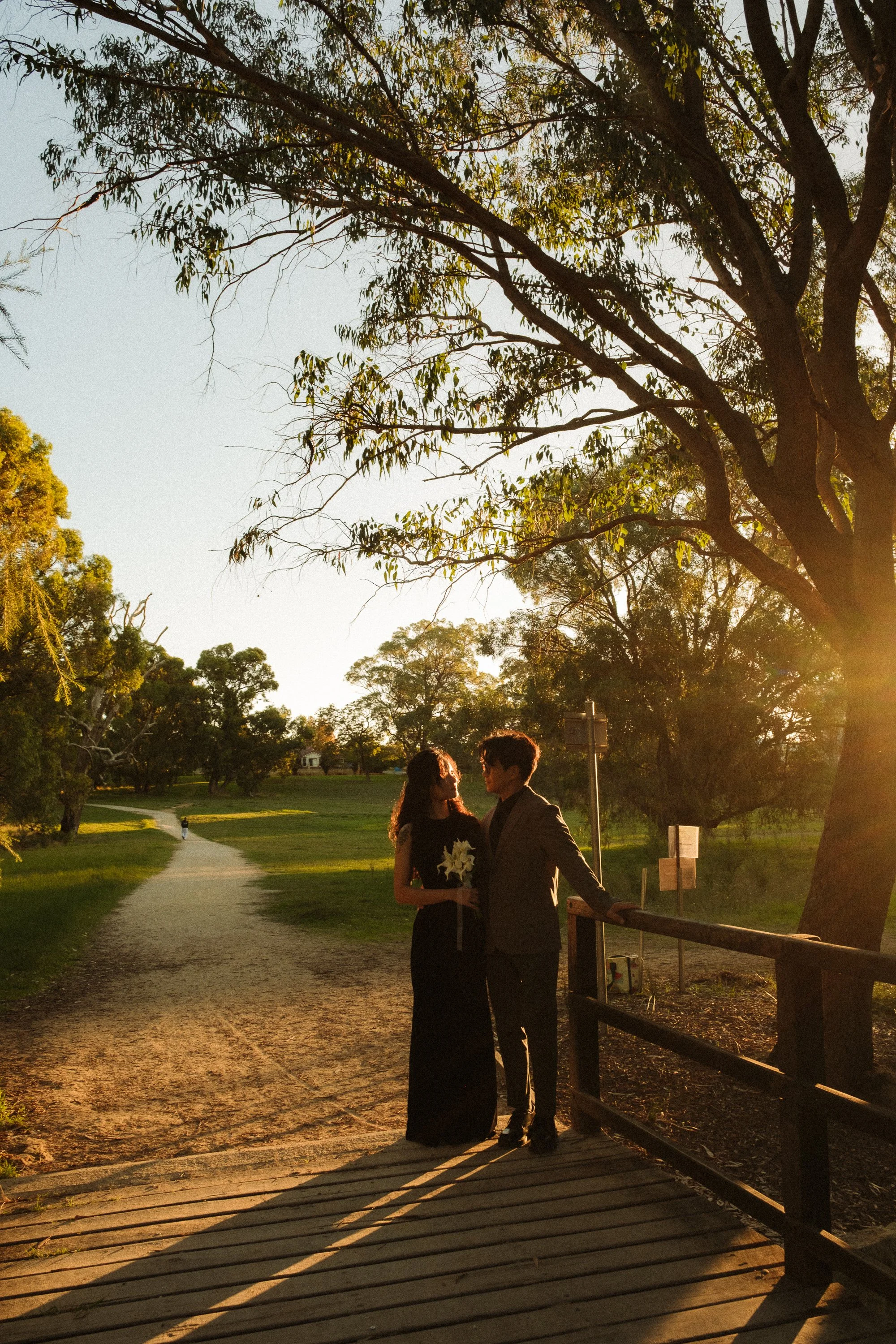 A couple dressed formally, standing on a small wooden bridge in a park during sunset, with trees and a pathway in the background.