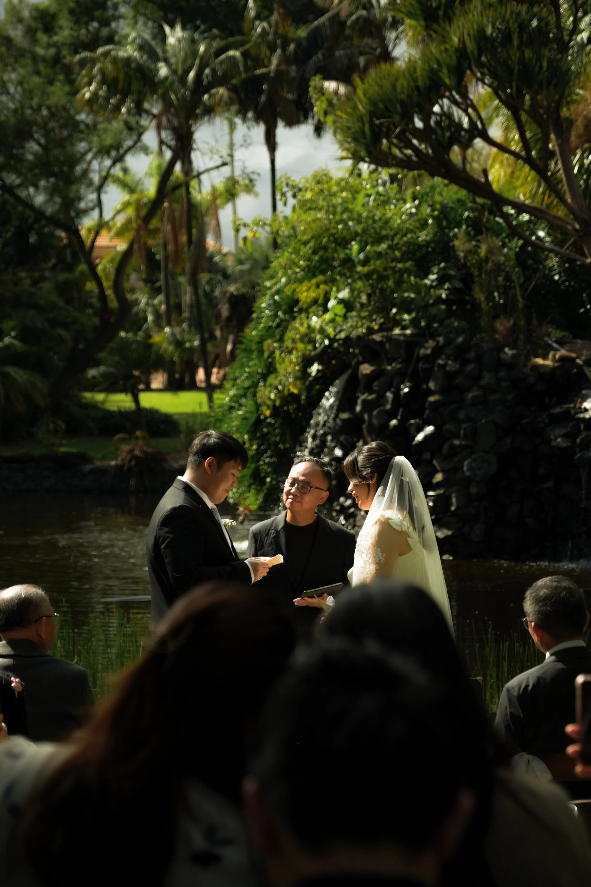 A wedding ceremony taking place outdoors by a pond, with three people standing and a crowd watching. The couple, dressed in wedding attire, facing each other with an officiant in front of them.