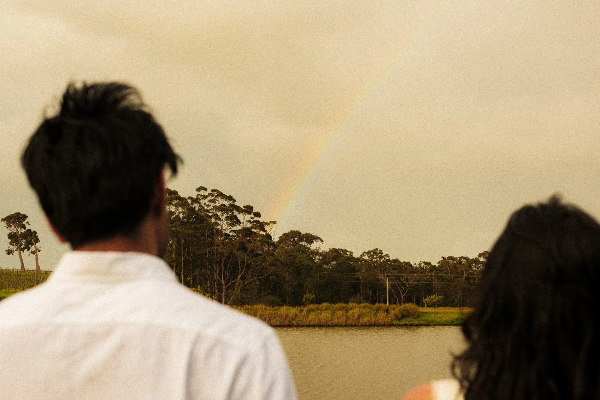 A man and a woman standing near a lake, looking at a rainbow in the sky over a wooded area.