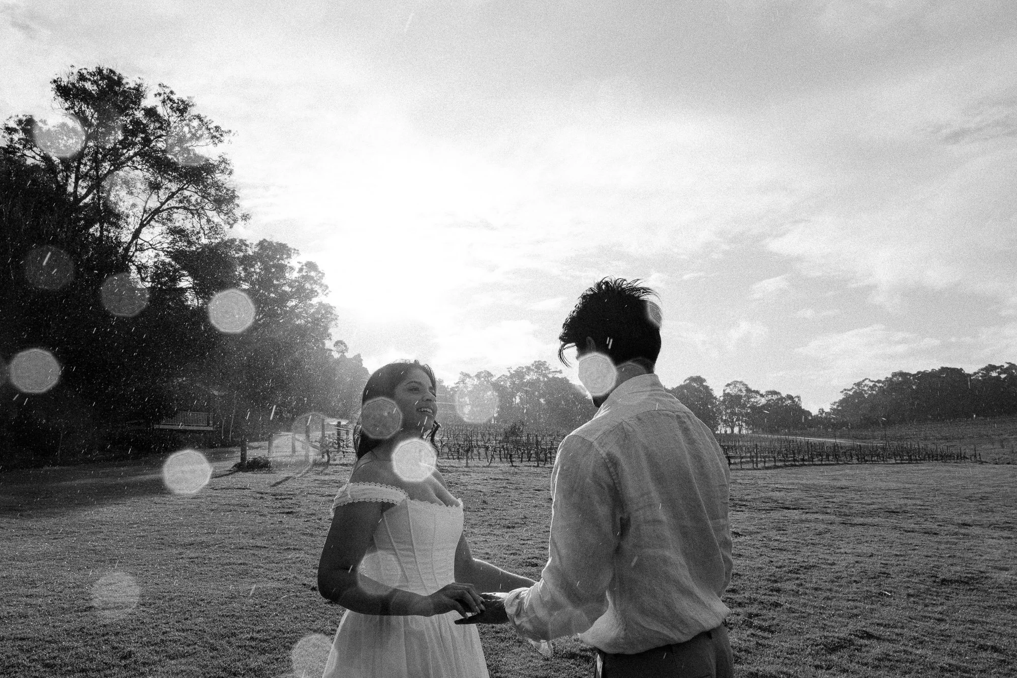 A black-and-white photo of a couple holding hands outside on a grassy field with trees and a fence in the background, as the sun sets behind them.