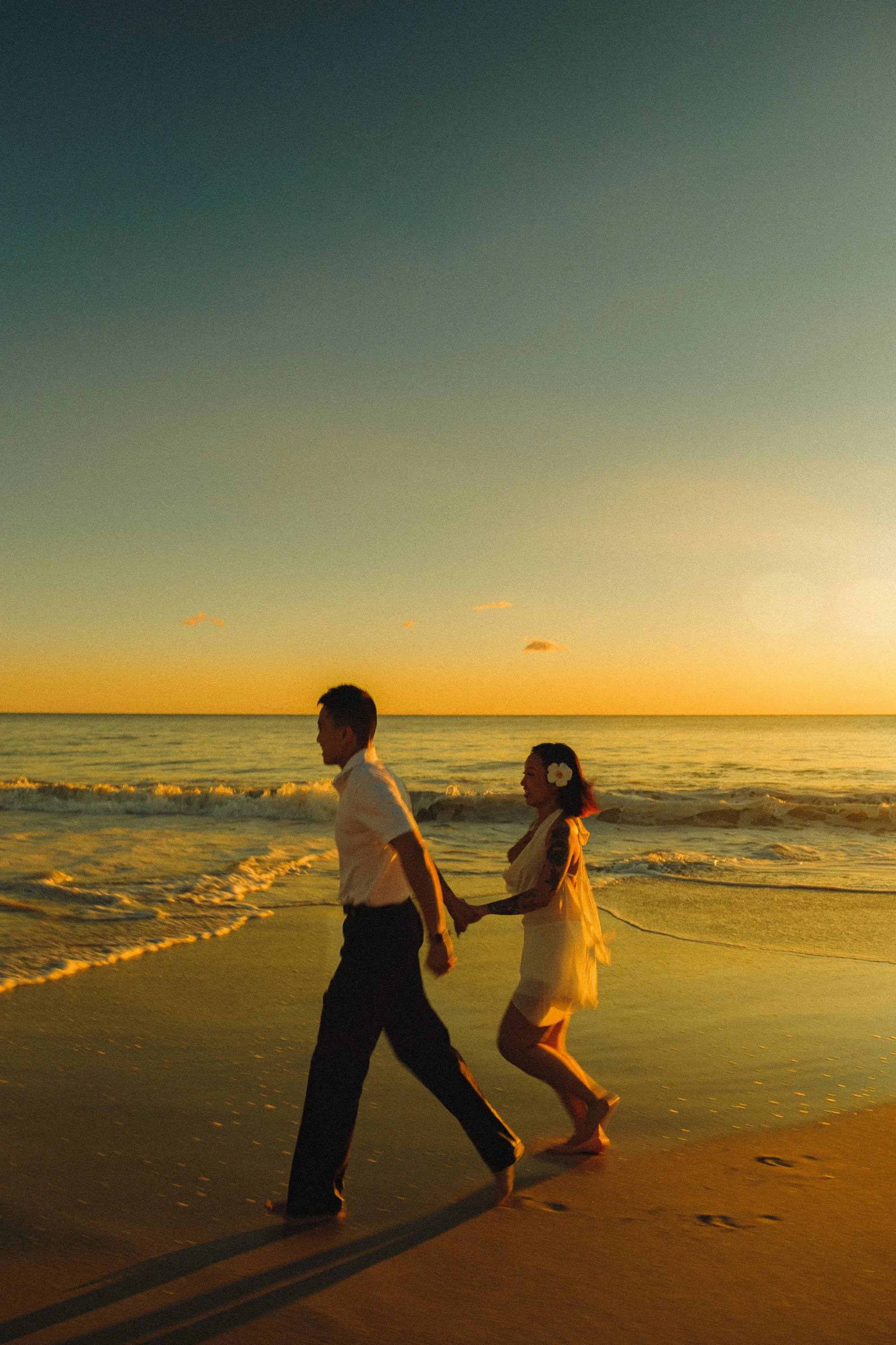 A couple holding hands and walking along the beach at sunset, with the man in a white shirt and dark pants, and the woman in a white dress with a flower in her hair.