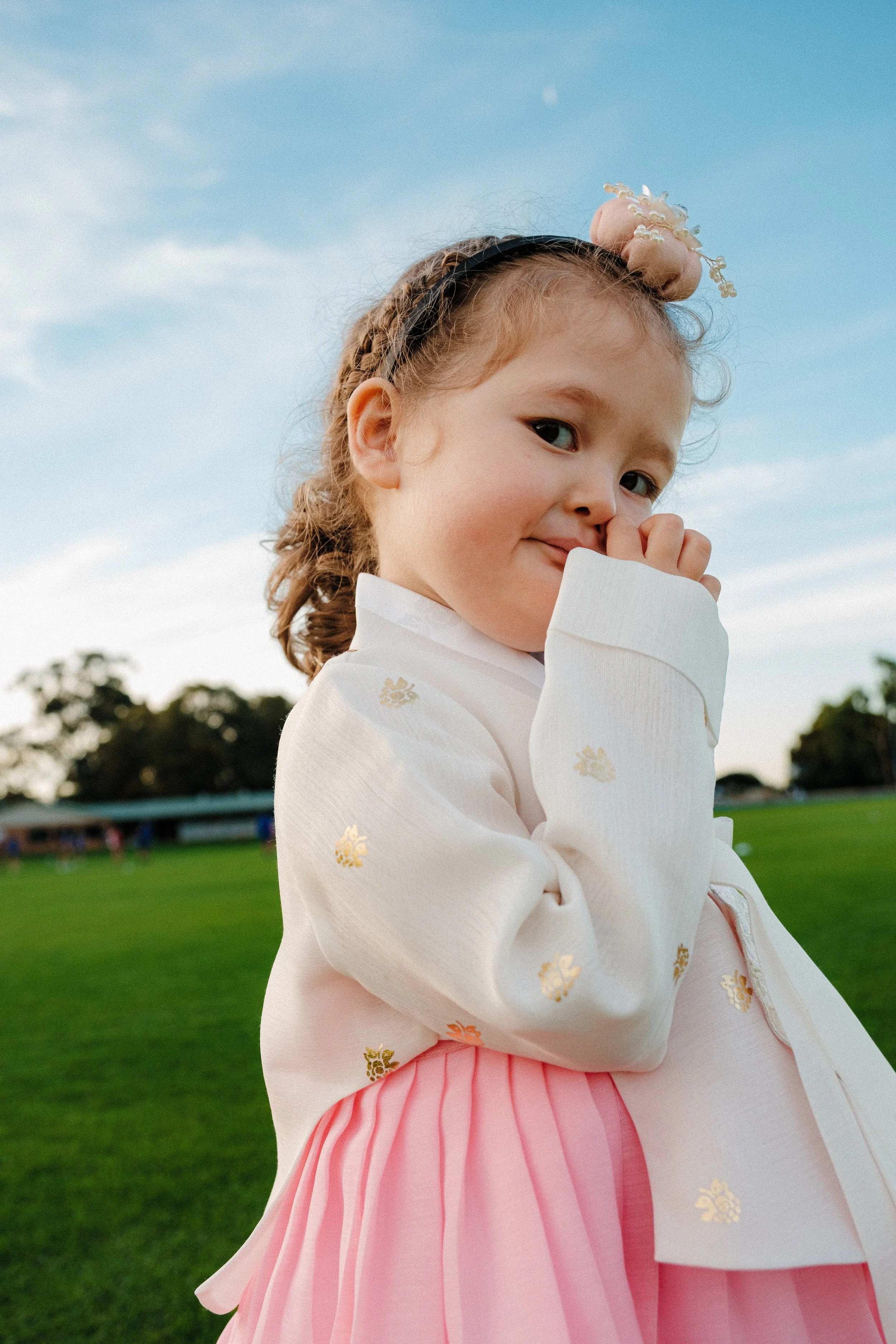 A young girl with curly hair wearing a white top with gold floral embroidery and a pink pleated skirt, standing outdoors on a grassy field with trees in the distance and a blue sky overhead.