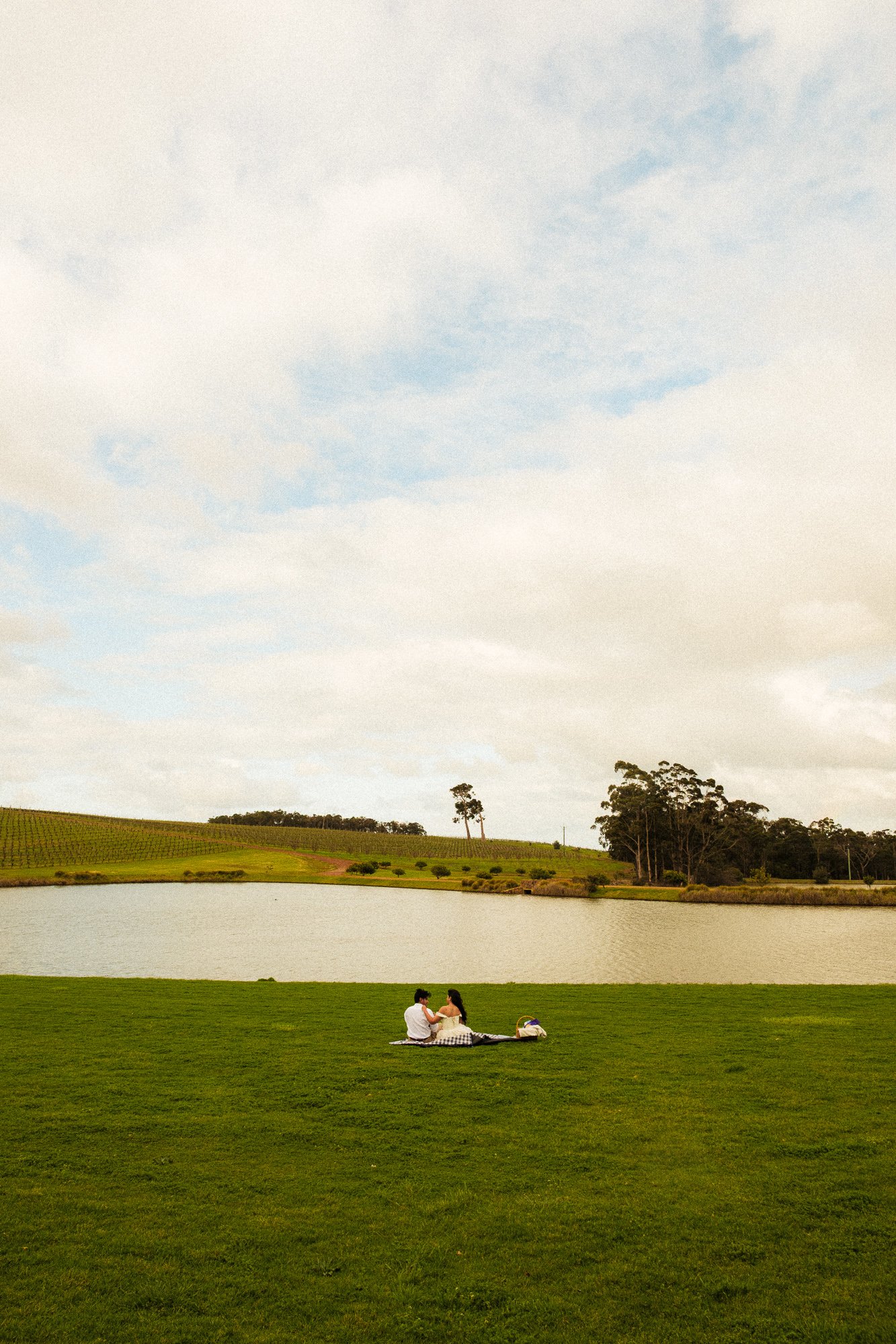 Two people sitting on a blanket near a lake, with green grass, trees, and a partly cloudy sky in the background.